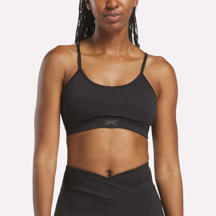 A woman models the Reebok Women's Yoga Strappy Bra, a black, medium-support, sweat-wicking bra with thin straps and a small front logo, paired with high-waisted black leggings against a plain light background (mid-torso up).