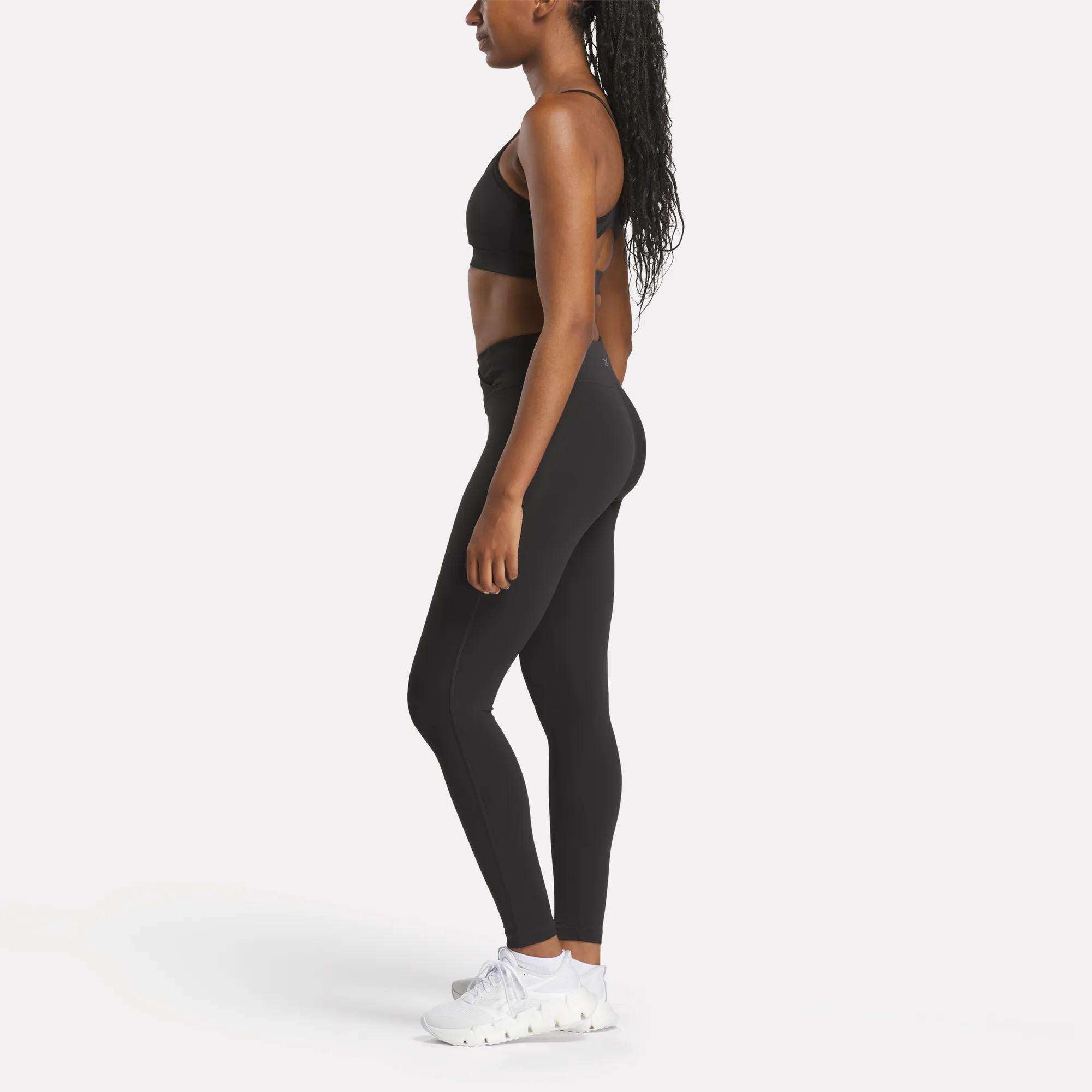 A woman stands in profile against a white background, wearing the Reebok Women's Yoga Strappy Bra in black, paired with black leggings and white athletic shoes. Her long, curly hair is worn down.