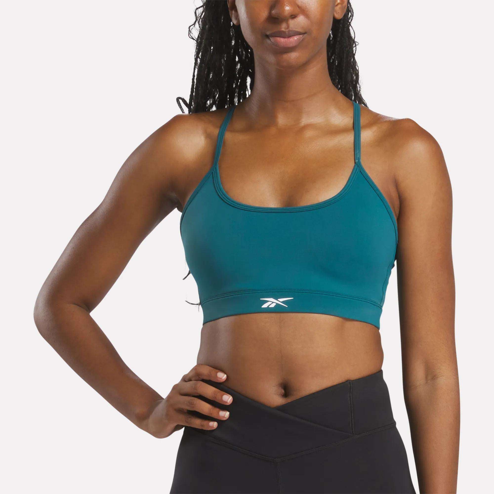 A woman wears the Reebok Women's Core Strappy Bra - Medium Support in teal with thin straps and a white logo, ideal for medium-impact workouts, paired with black athletic leggings.