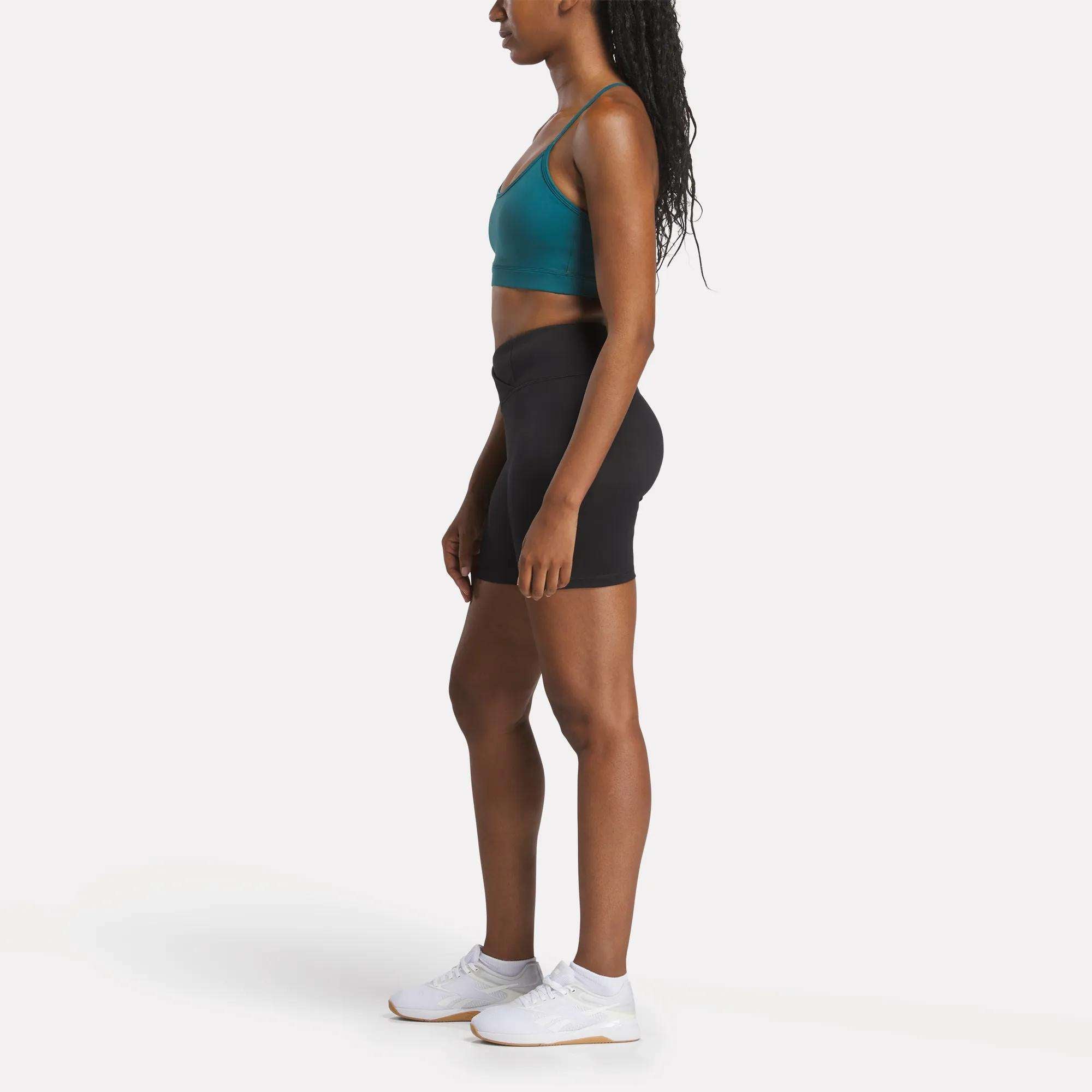 A woman stands in profile against a white background, wearing the Reebok Women's Core Strappy Bra - Medium Support in teal, paired with black high-waisted shorts and white sneakers.