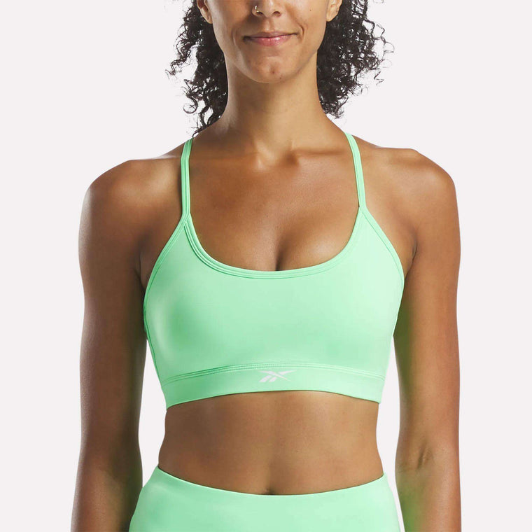 A woman models the Reebok Women's Core Strappy Bra - Medium Support in light green, featuring thin straps and a small chest logo. The quick-drying bra is ideal for medium-impact workouts and stands out against a plain white background.