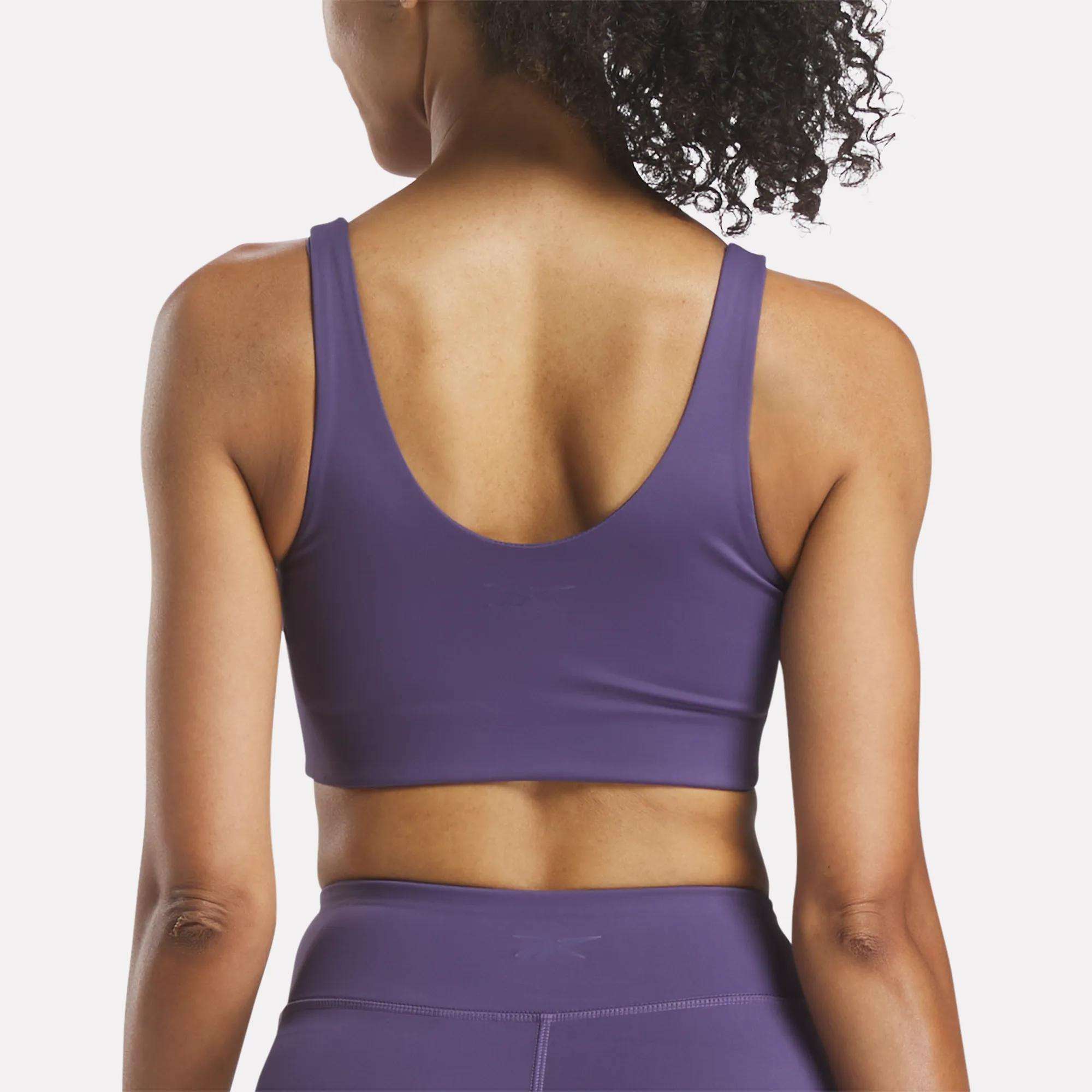 A woman with curly hair, seen from behind, wears a purple Reebok Women's Yoga V-Neck Bra and matching leggings against a plain light background—designed for studio workouts and medium-impact support.