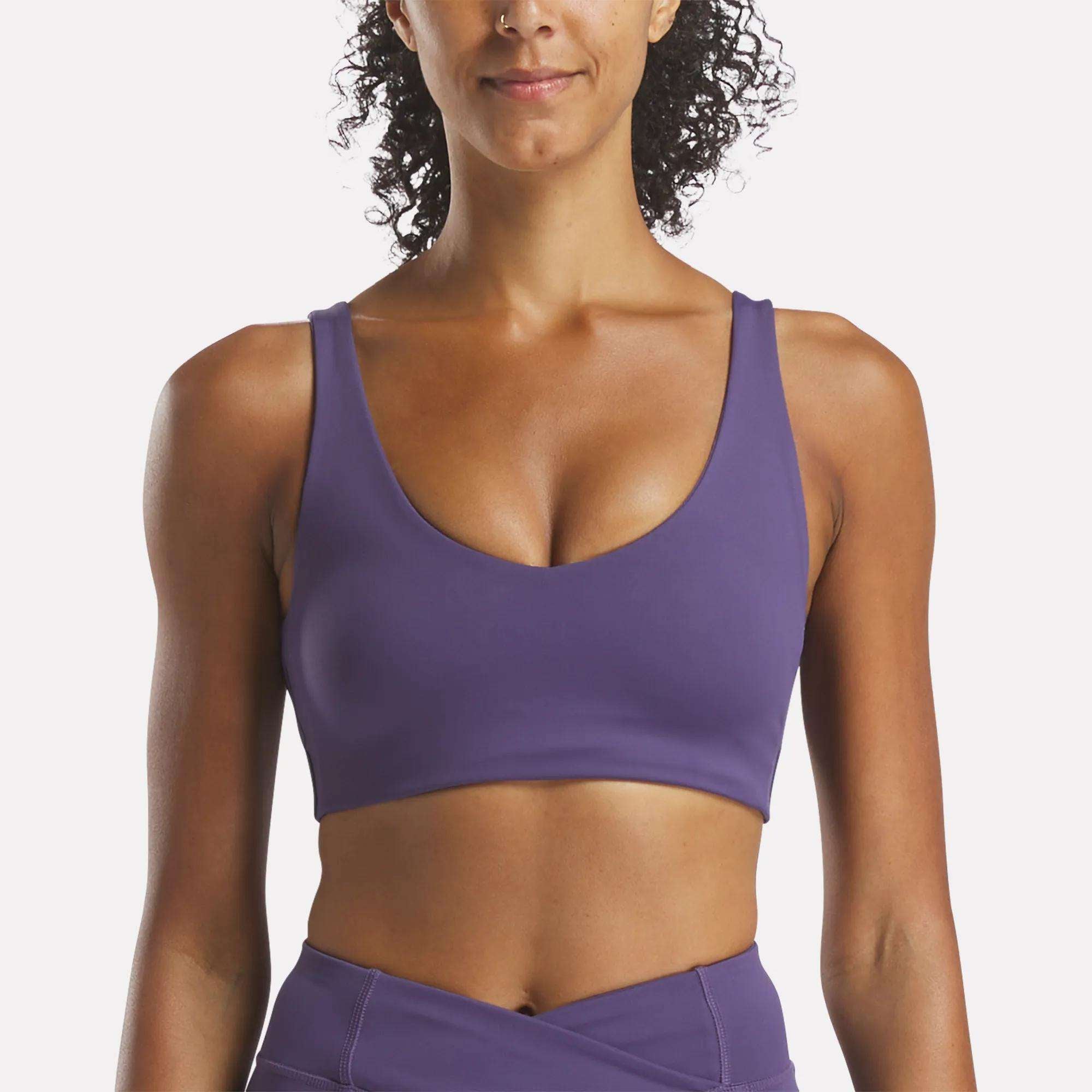 A woman wears the Reebok Women's Yoga V-Neck Bra in purple with matching high-waisted leggings, styled for medium-impact support—perfect for studio workouts—shown from the waist up against a plain white background.