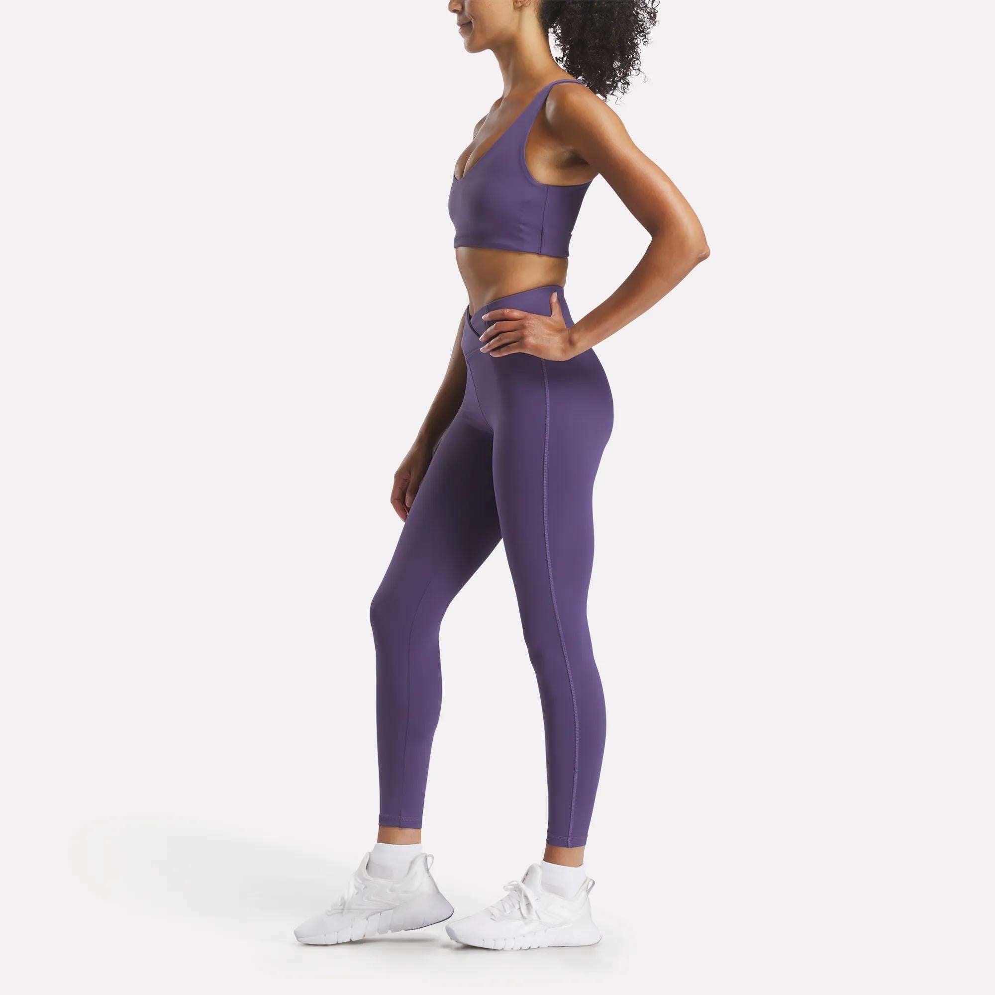 A woman wears the Reebok Women's Yoga V-Neck Bra, a purple sports bra designed for medium-impact support during studio workouts.