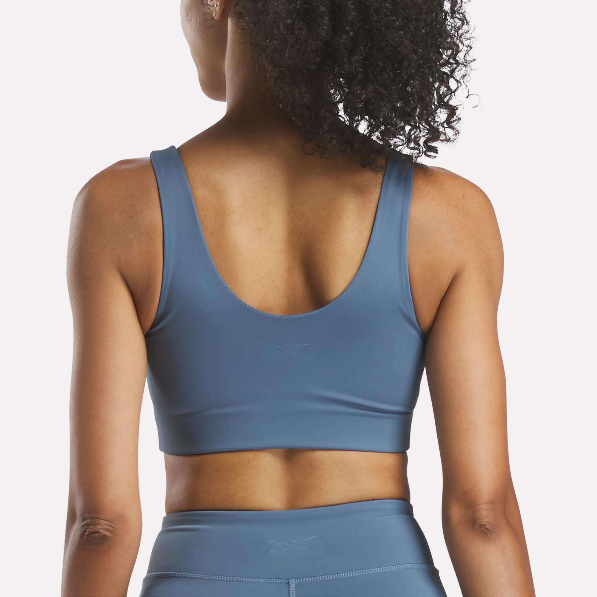 A woman with curly hair, seen from behind, wears the Reebok Women's Yoga V-Neck Bra in blue with matching high-waisted leggings against a plain light background—perfect for studio workouts.