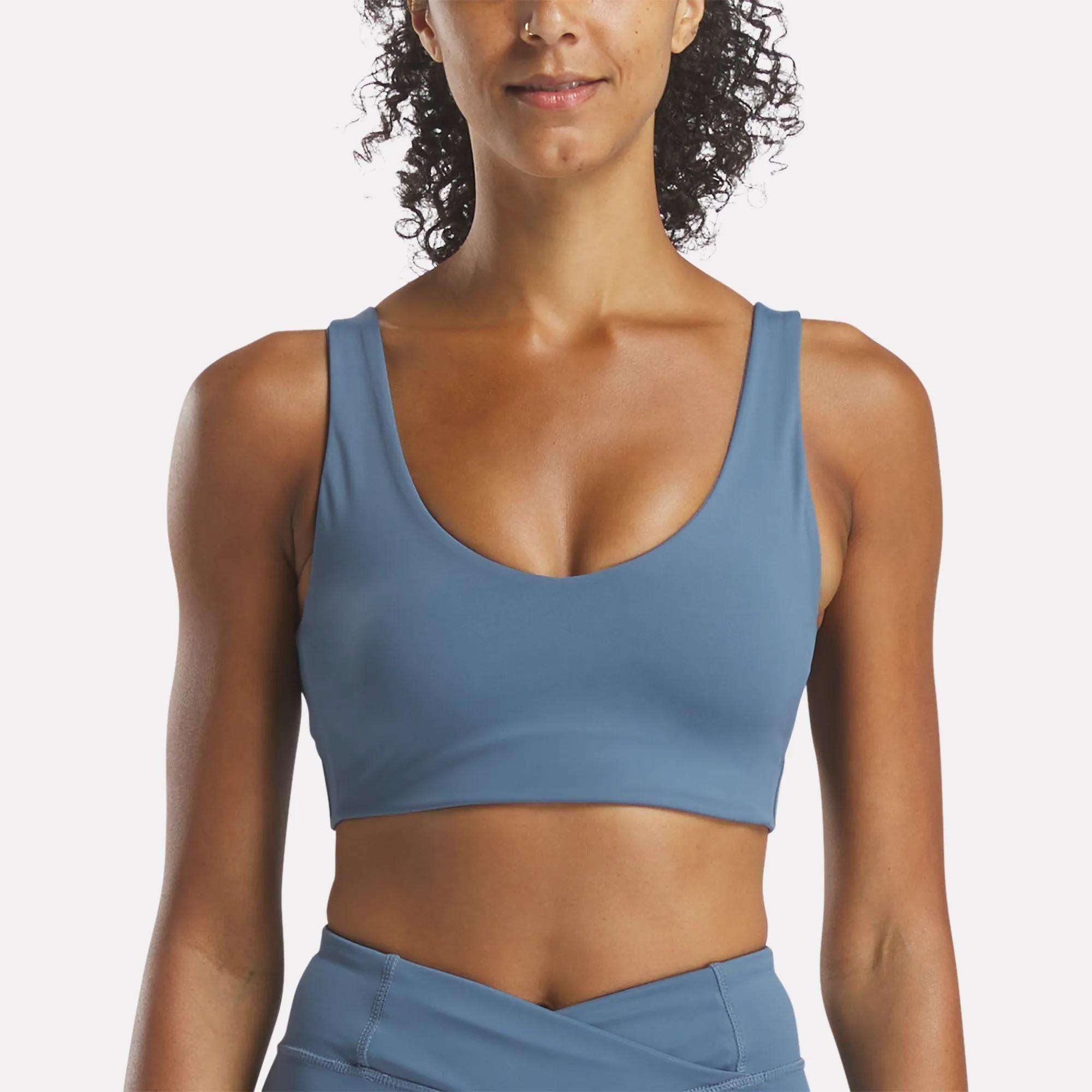 A woman with curly hair models the Reebok Women's Yoga V-Neck Bra and matching high-waisted leggings in blue, shown from shoulders to hips against a plain white background—ideal for studio workouts.