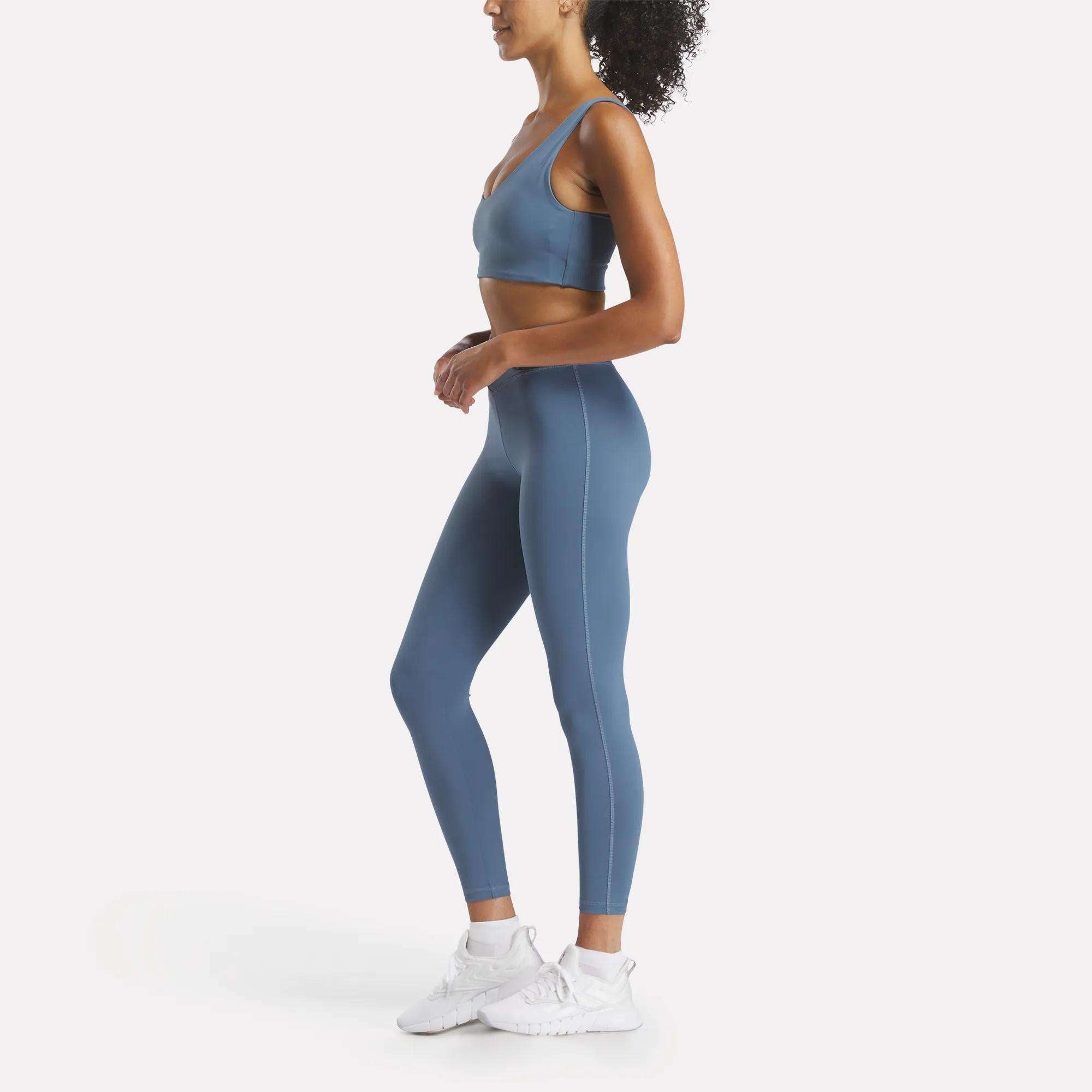 Woman in a blue Reebok Women's Yoga V-Neck Bra with medium support and matching high-waisted leggings stands in profile against a white background, ready for studio workouts in white sneakers.