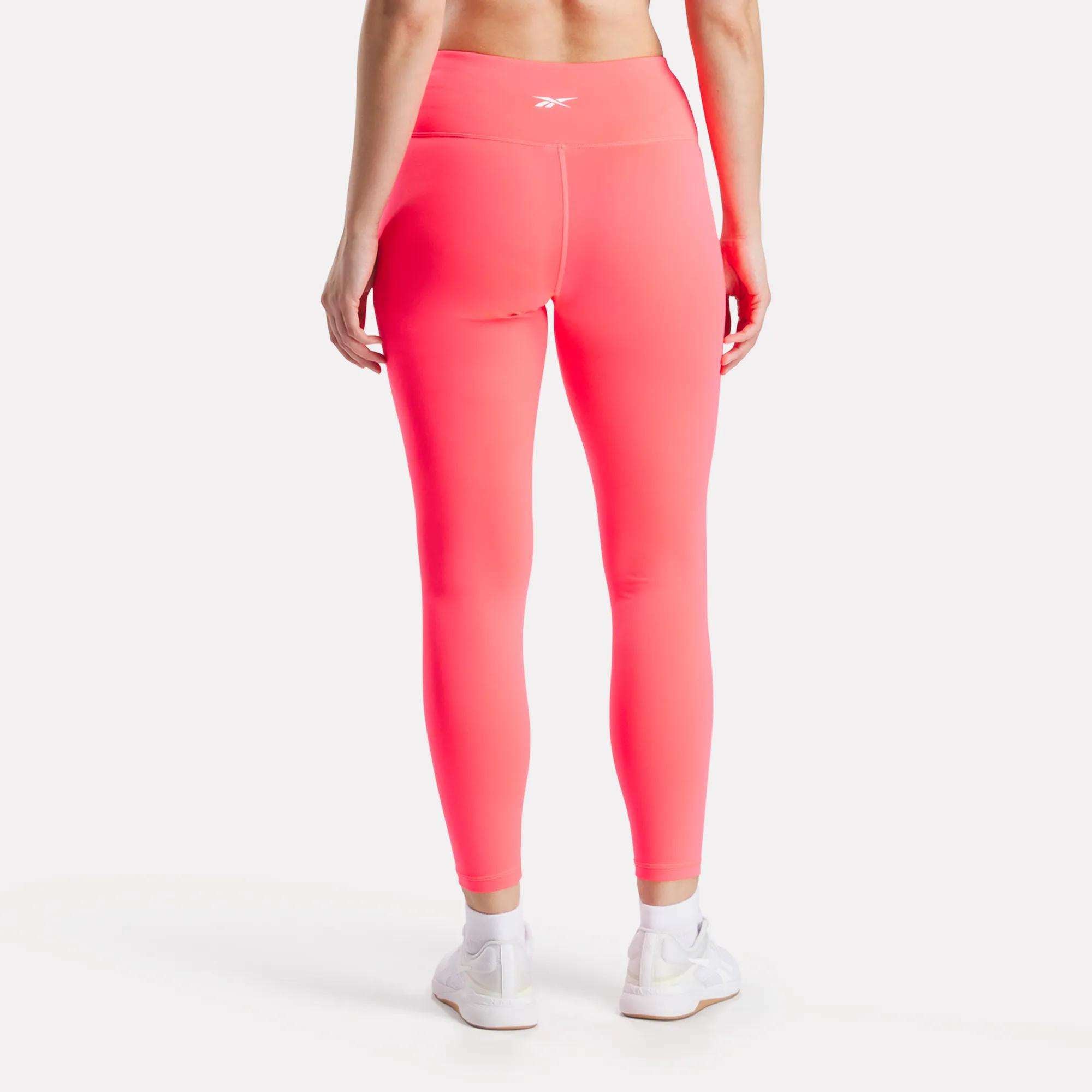A woman is shown from the back, wearing Reebok Women's Core High-Rise Leggings in bright pink, made with RBK-DRY fabric, and white sneakers against a plain white background.