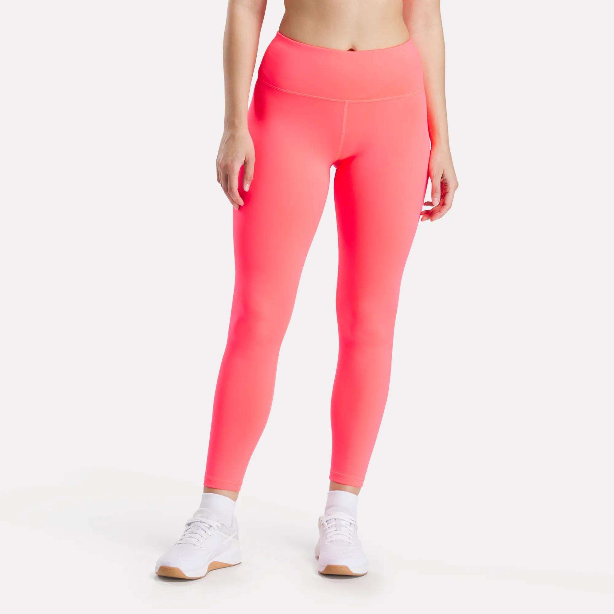 A woman models Reebok Women's Core High-Rise Leggings in bright pink, paired with white sneakers, standing against a plain white background; most of her upper body is out of frame.