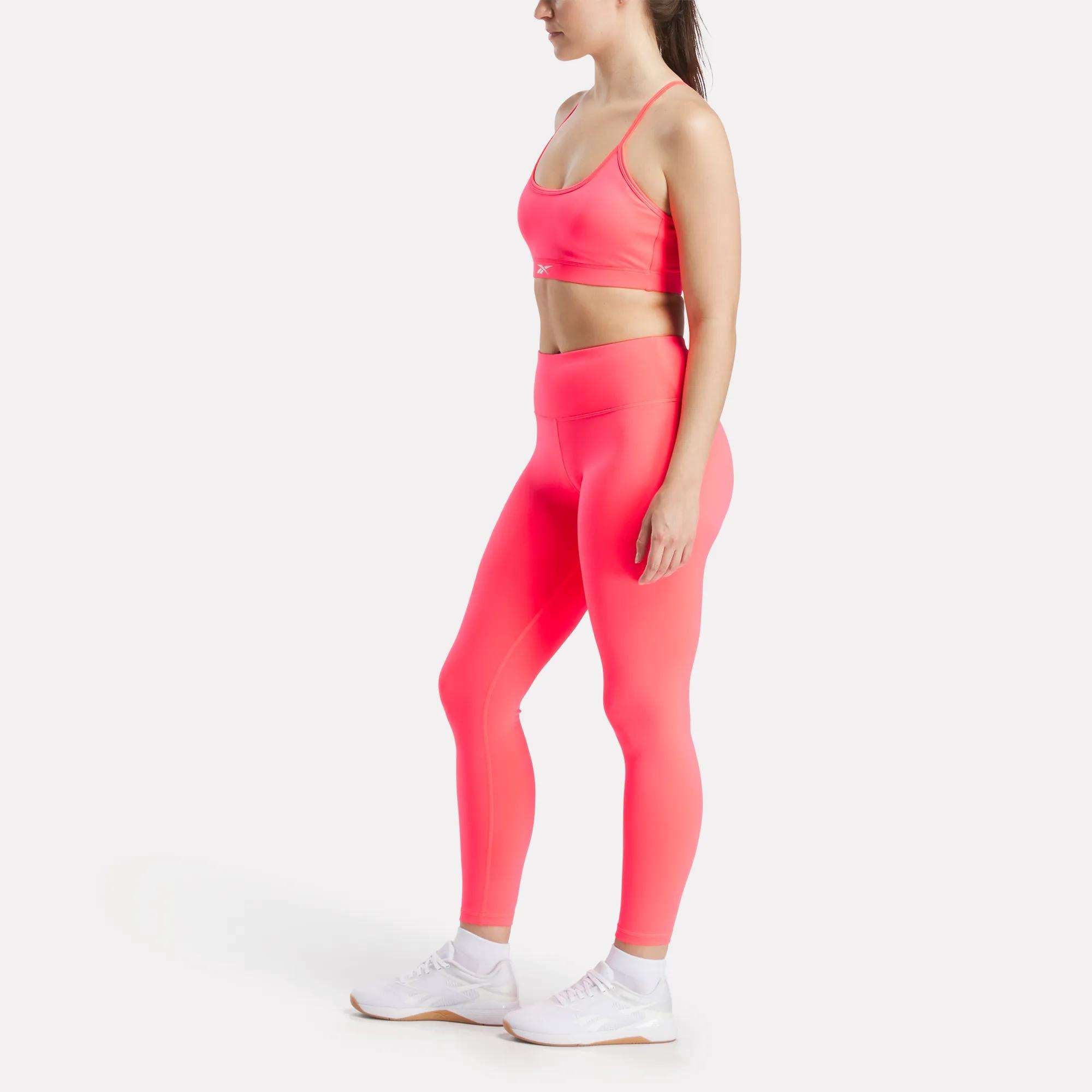 A woman stands in profile wearing Reebok's Women's Core High-Rise Leggings and a matching sports bra, both in bright pink RBK-DRY fabric, paired with white sneakers against a plain light background.