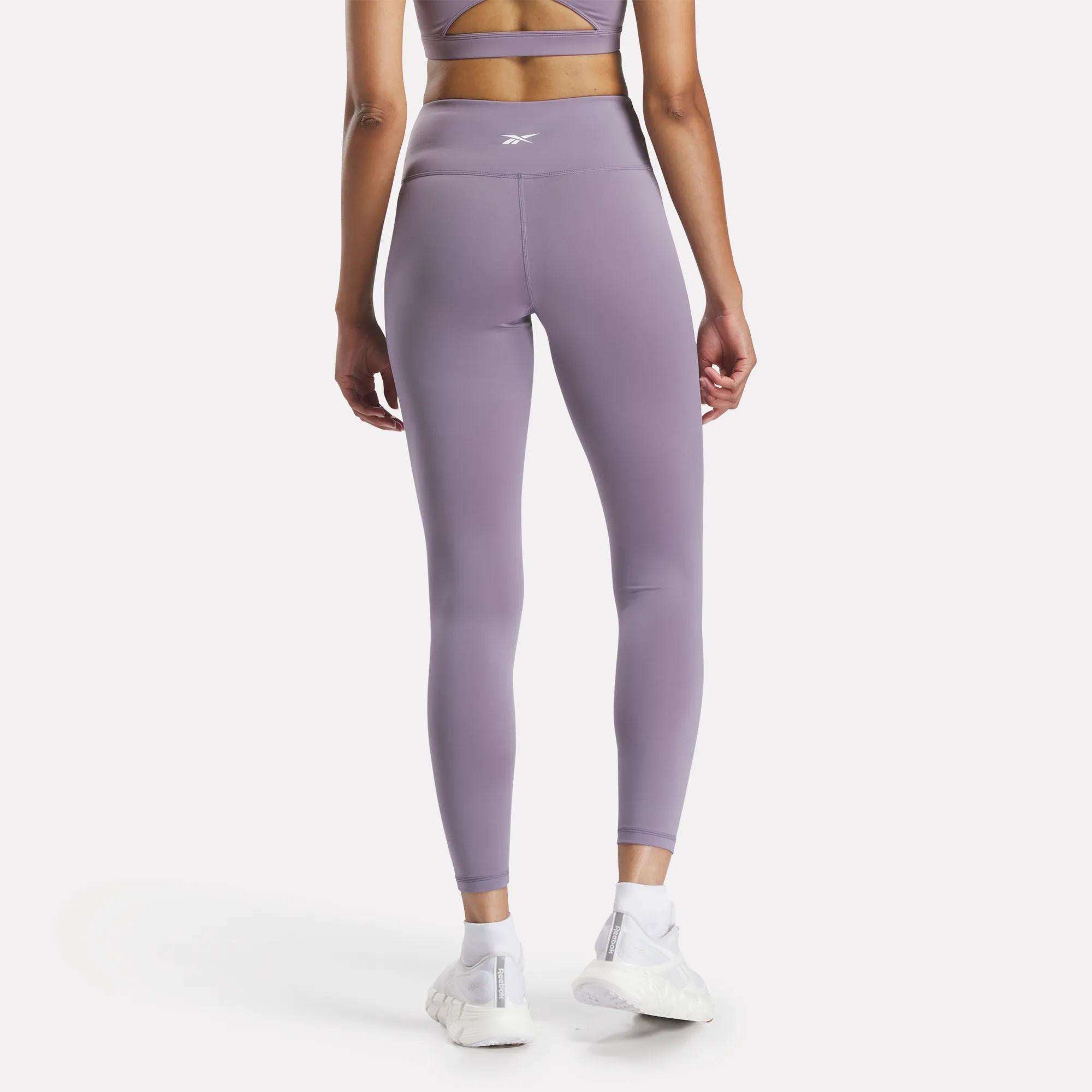 A woman faces away in Reebok Women's Core High-Rise Leggings and a lavender sports bra, both made from RBK-DRY fabric, with white sneakers. She stands against a light, plain background.