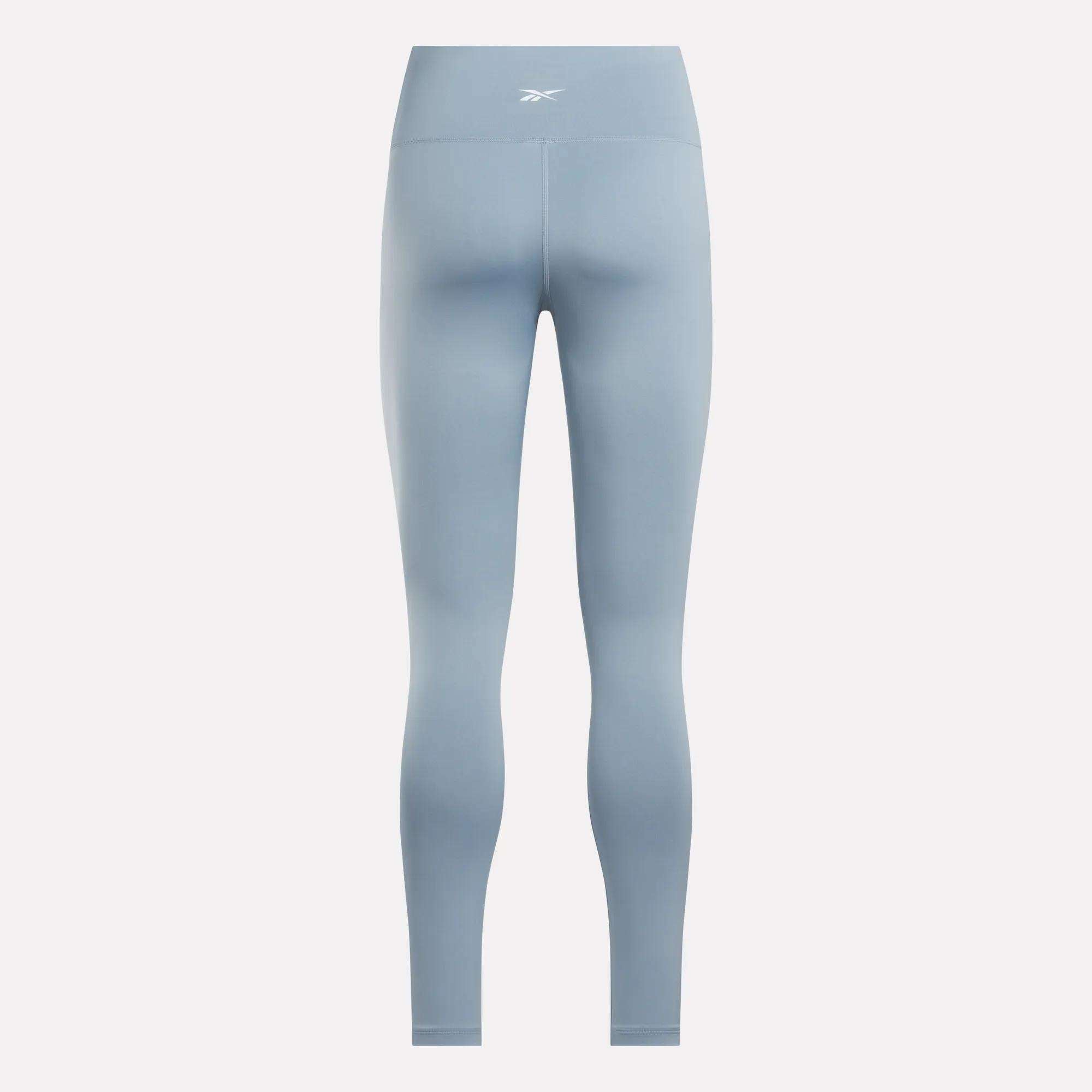 The Reebok Women's Core High-Rise Leggings in light blue feature a small white logo on the back waistband, RBK-DRY fabric for moisture wicking, a smooth seamless design, and full-length legs.