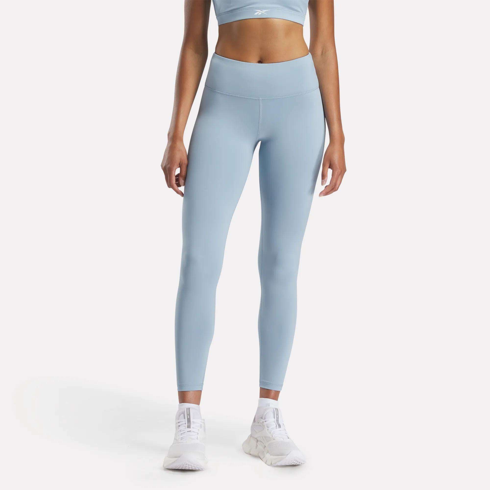 A person wears Reebok Women's Core High-Rise Leggings in light blue, paired with a matching sports bra and white athletic shoes, standing against a plain white background. The cropped photo shows the model from shoulders to feet.