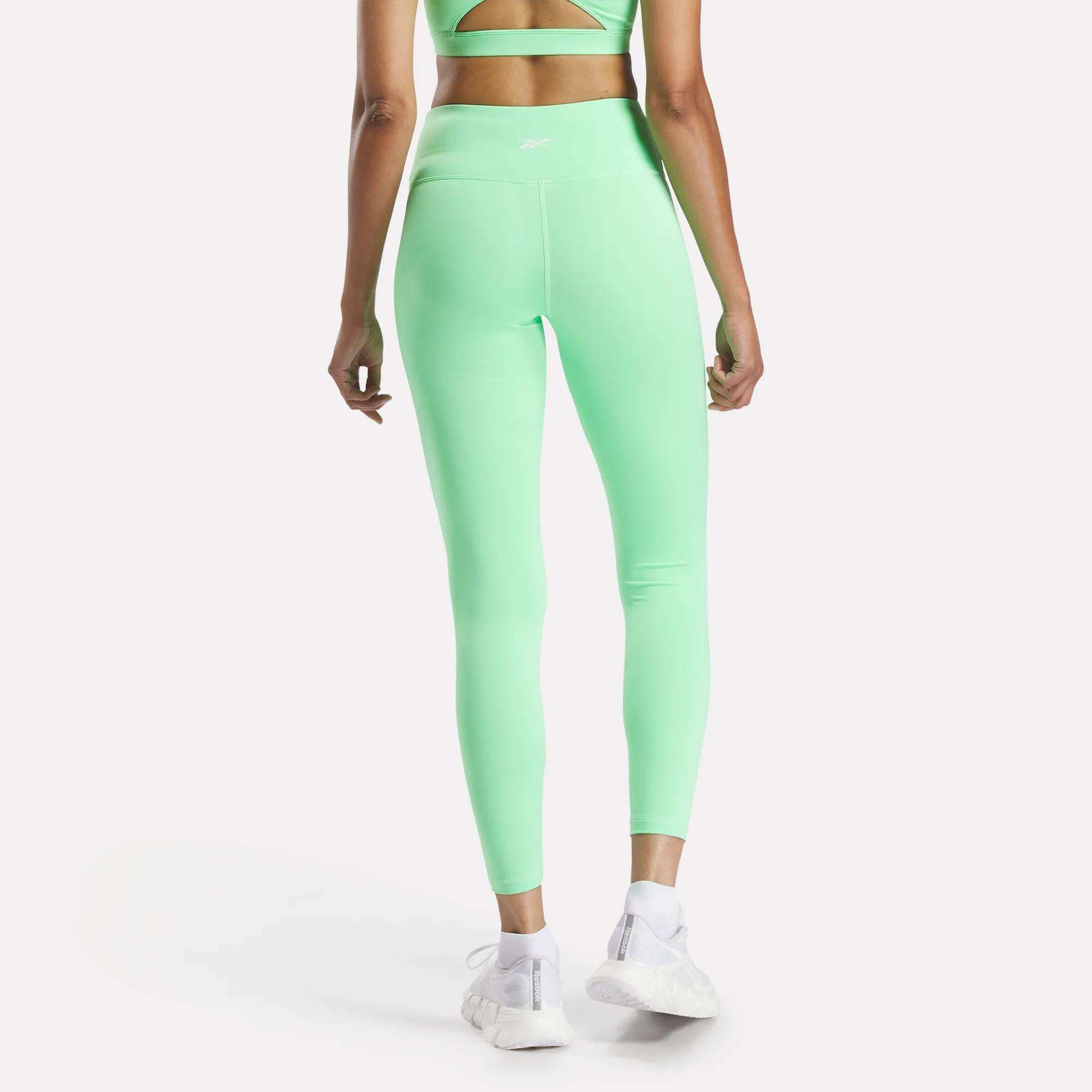 A woman faces away wearing Reebok Women's Core High-Rise Leggings in light green with a matching sports bra and white sneakers, standing against a plain white background. Both pieces feature moisture-wicking RBK-DRY fabric.