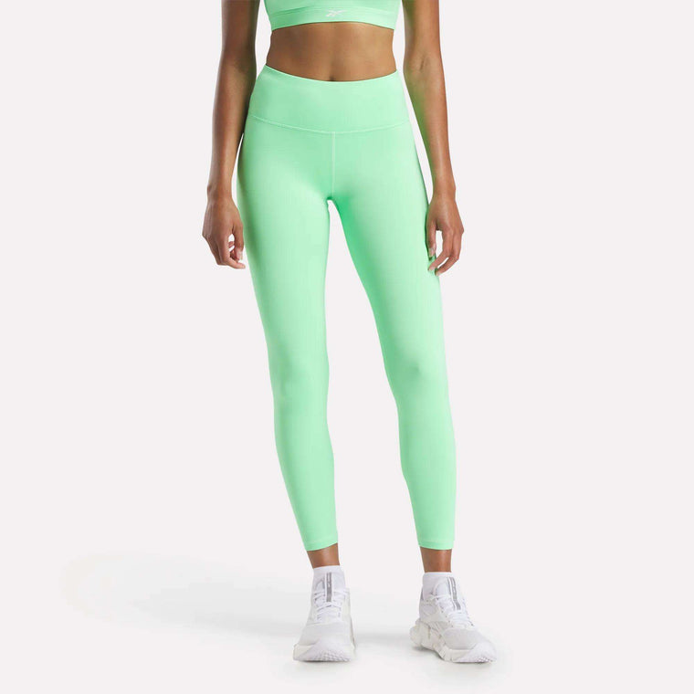 A woman models Reebok's Women's Core High-Rise Leggings in neon green with a matching sports bra, both crafted from RBK-DRY fabric. She pairs them with white athletic shoes, standing against a plain white background.