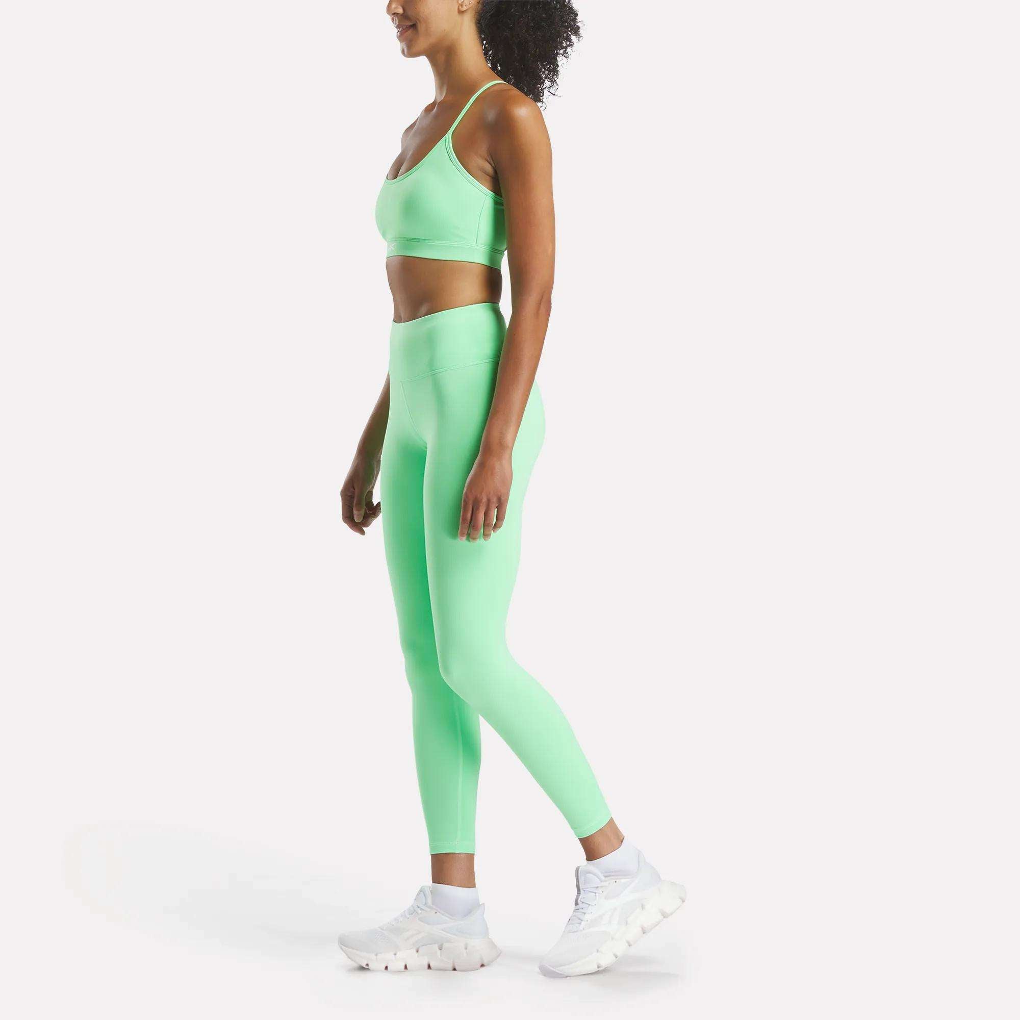 A woman wears Reebok Women's Core High-Rise Leggings in bright green with a matching sports bra and white sneakers, standing sideways against a plain white background with her curly hair pulled back.