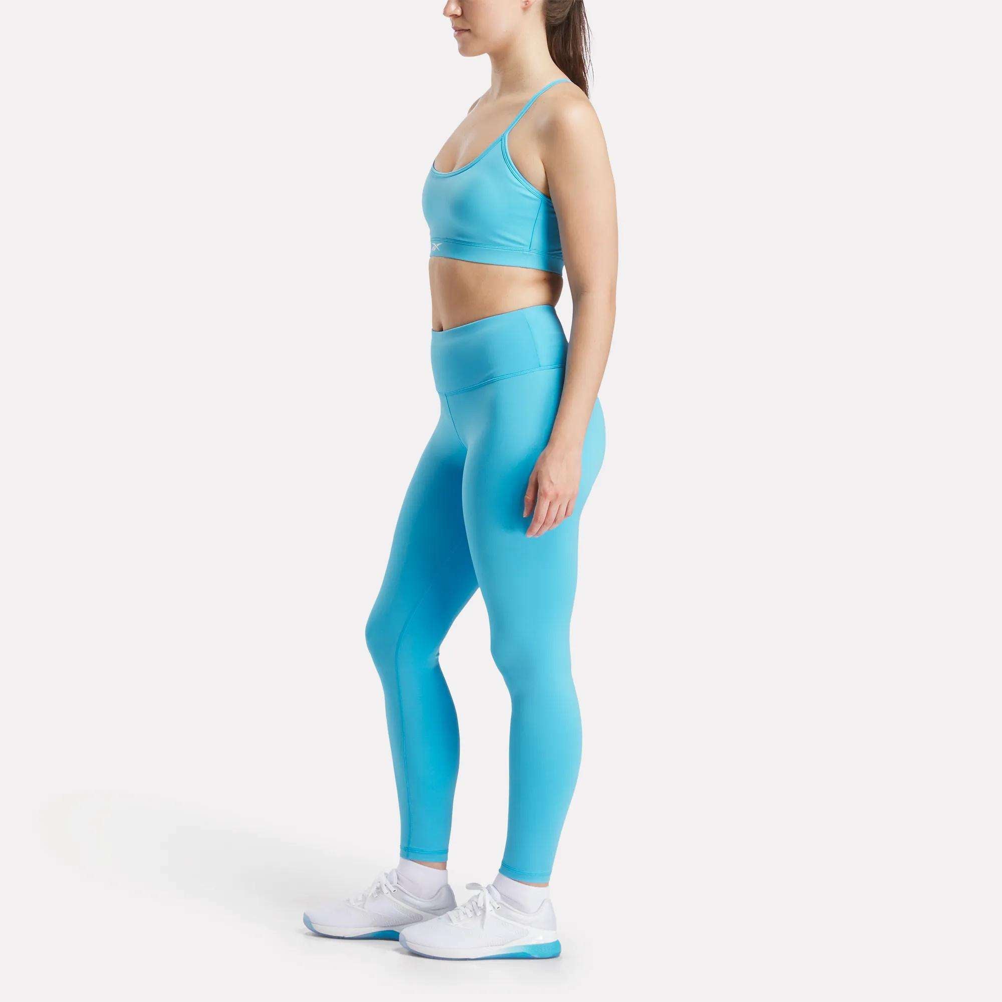 A woman stands in profile on a white background wearing Reebok Women’s Core High-Rise Leggings and a matching light blue sports bra, both made from RBK-DRY fabric, paired with white sneakers.