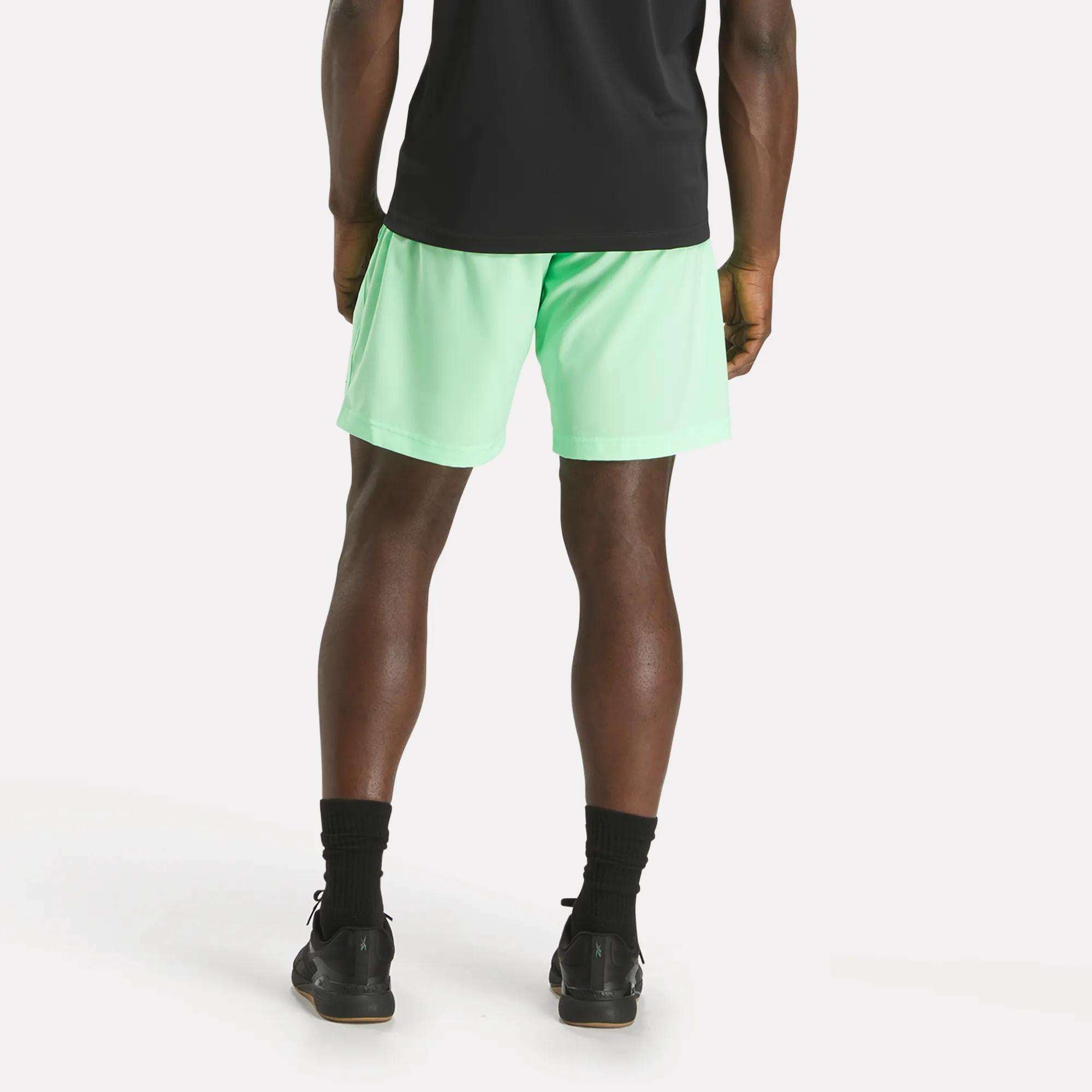 A person stands facing away from the camera, wearing a black shirt and mint green Reebok Men's Workout Ready Woven Shorts, paired with black socks and shoes, set against a plain white background.