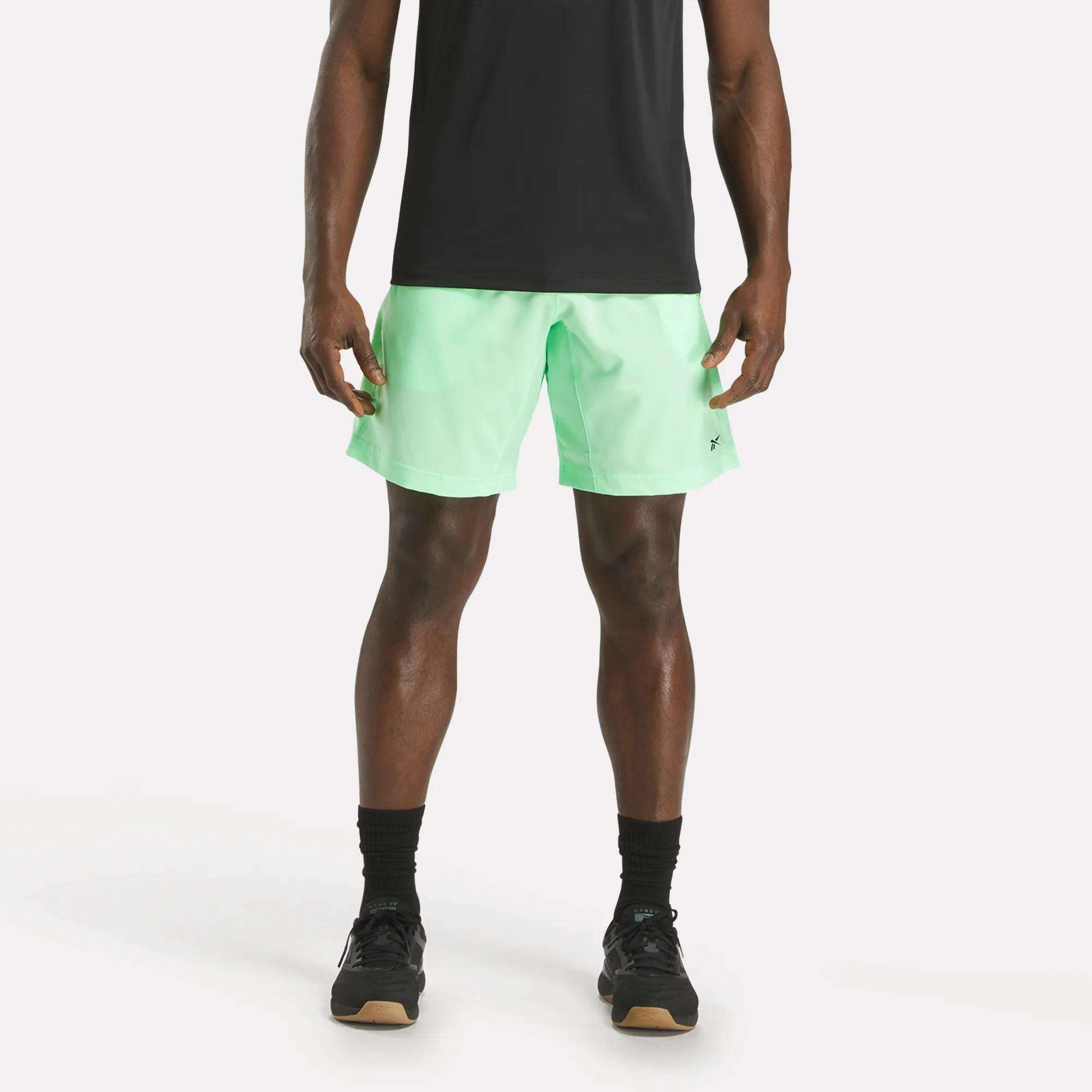 A man stands against a plain white background, wearing a black t-shirt, Reebok Men's Workout Ready Woven Shorts in light green, black socks, and black athletic shoes. His face is not visible in the image.