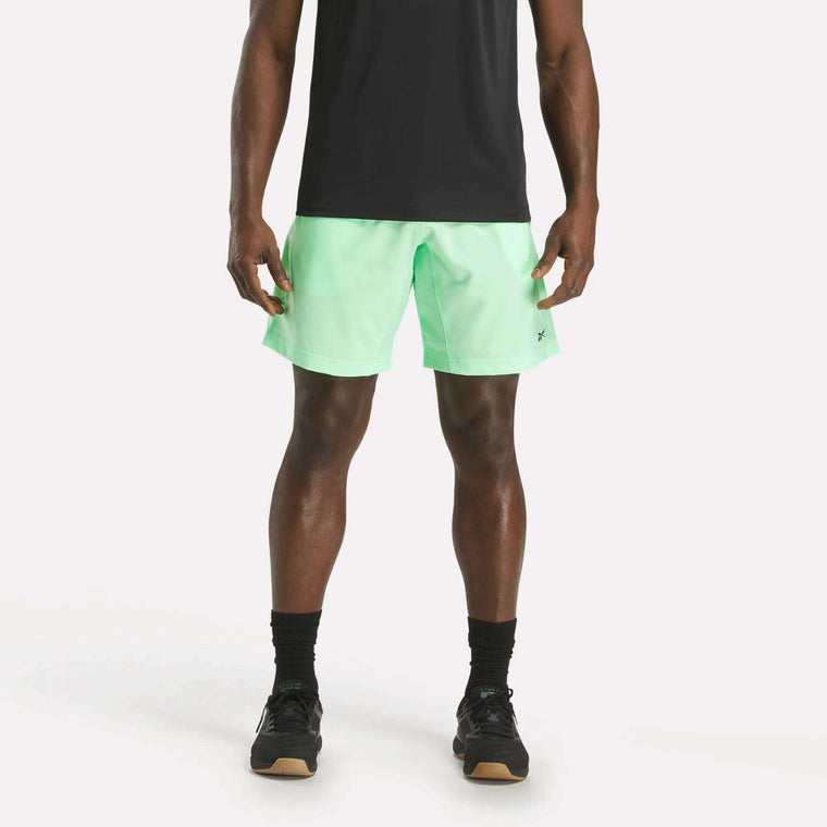 A man stands against a plain white background, wearing a black t-shirt, Reebok Men's Workout Ready Woven Shorts in light green, black socks, and black athletic shoes. His face is not visible in the image.