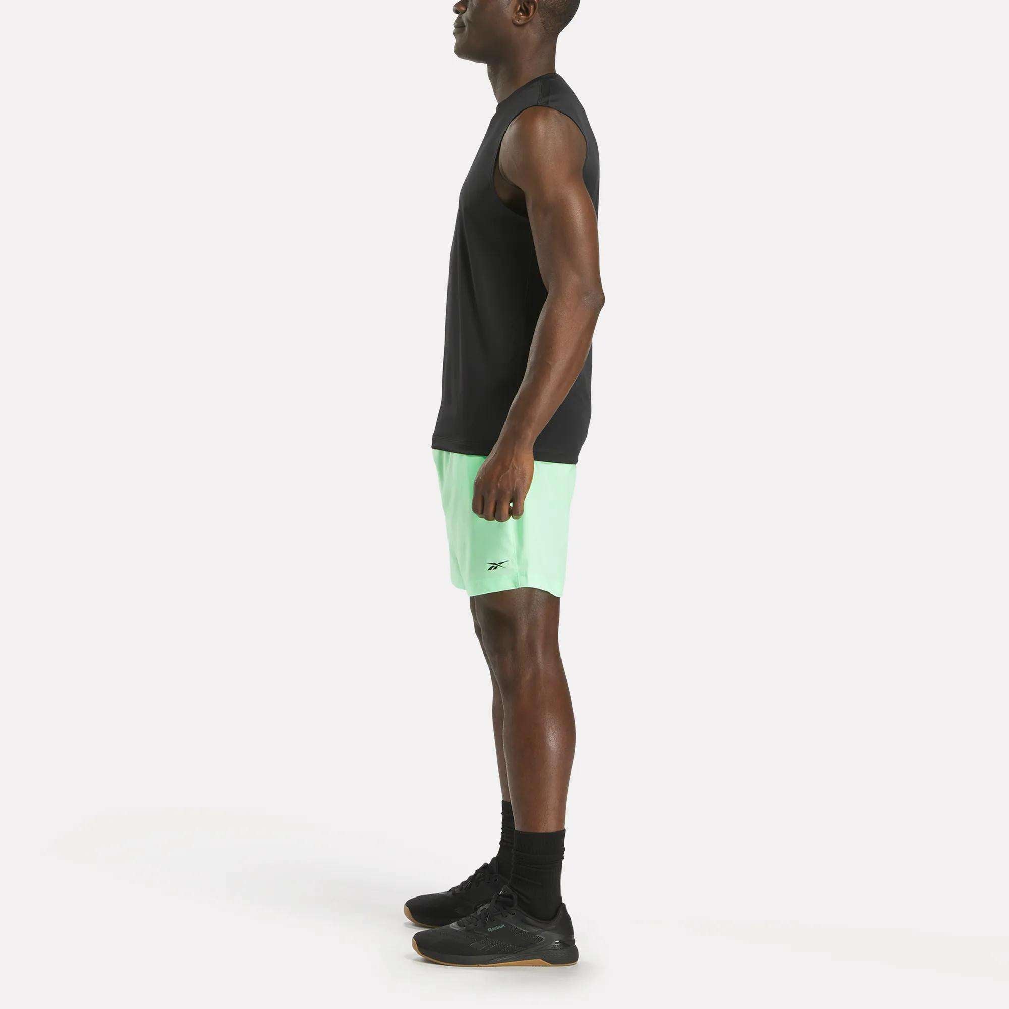 A man stands in profile against a plain white background wearing a sleeveless black shirt, black socks, black sneakers, and light green Reebok Men's Workout Ready Woven Shorts.