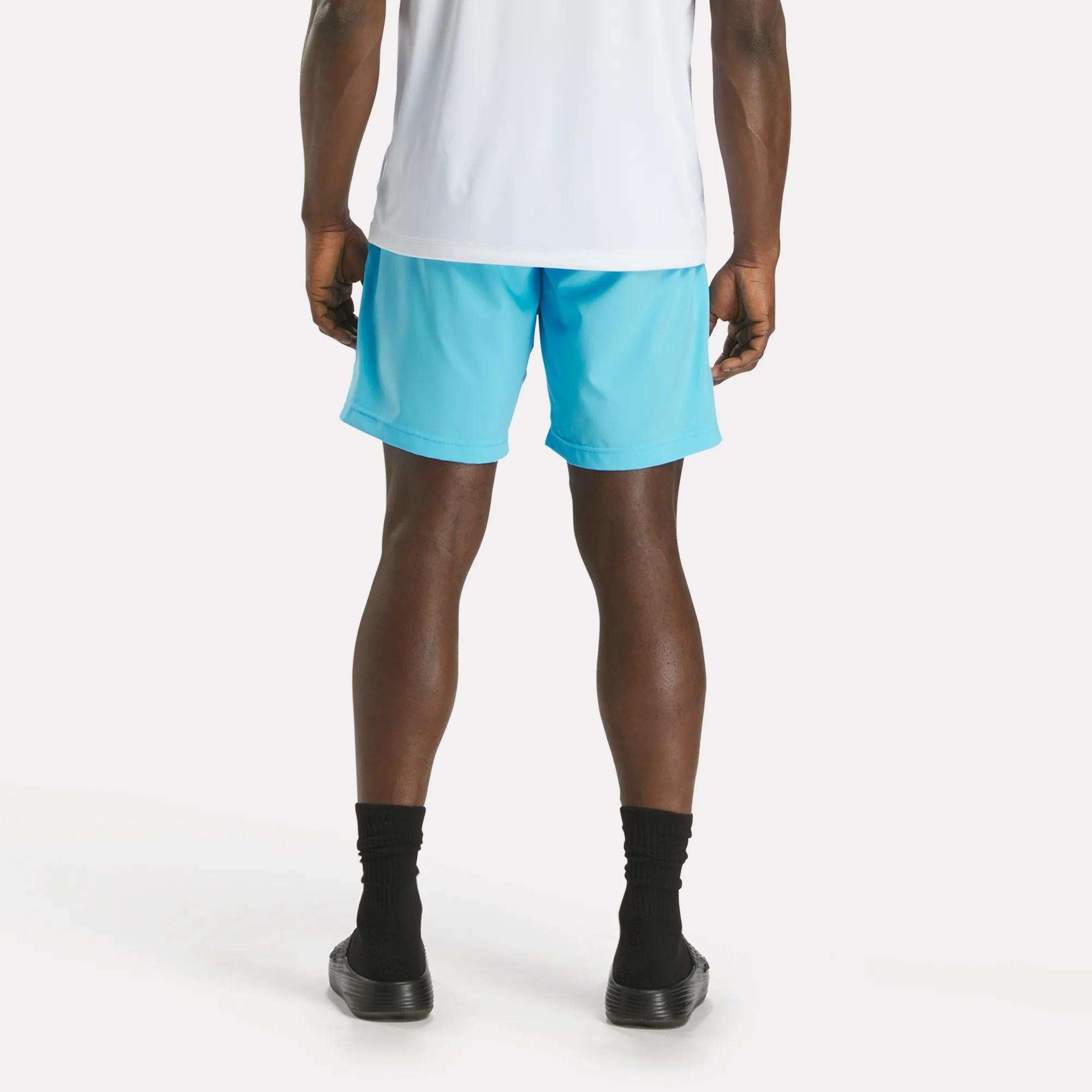 A person stands with their back to the camera, dressed in a white shirt and light blue Reebok Men's Workout Ready Woven Shorts, paired with black socks and black slip-on shoes against a plain white background.