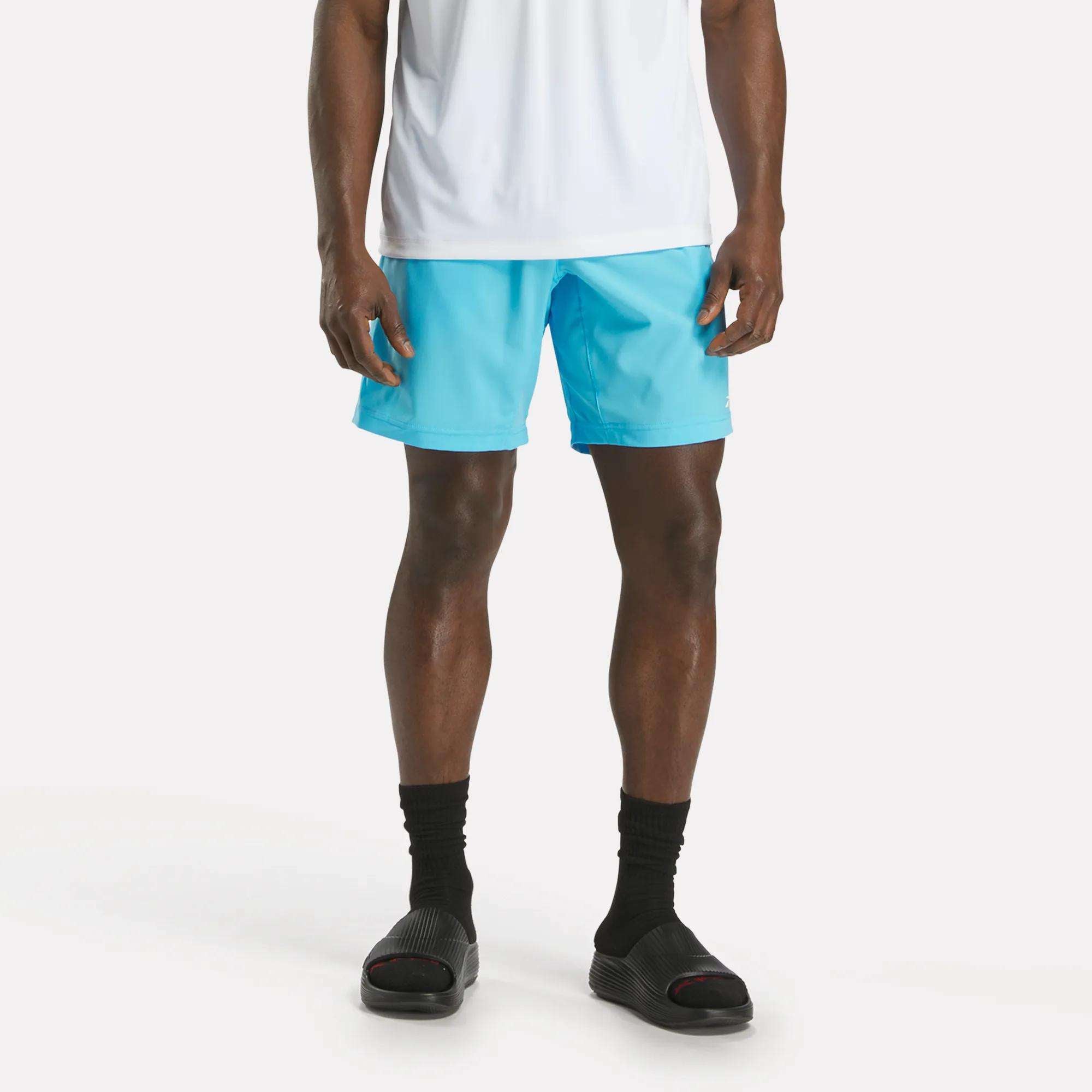 A person stands against a plain white background wearing a white shirt, black socks, black slide sandals, and bright blue Reebok Men's Workout Ready Woven Shorts. The person's upper body is partially out of frame.
