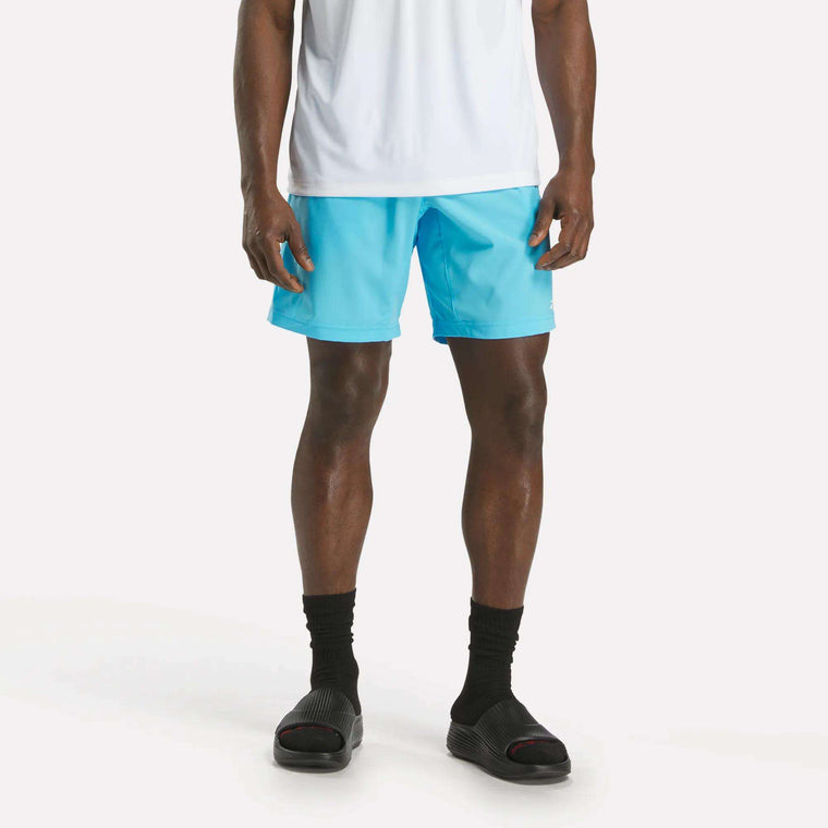 A person stands against a plain white background wearing a white shirt, black socks, black slide sandals, and bright blue Reebok Men's Workout Ready Woven Shorts. The person's upper body is partially out of frame.