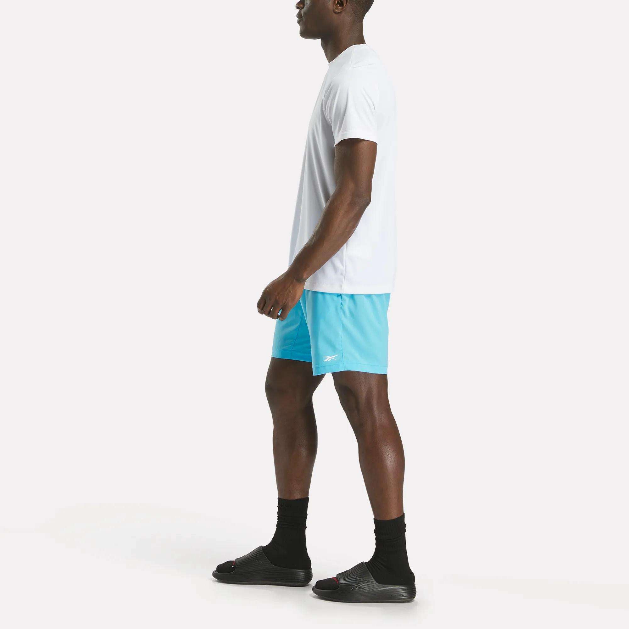 A man stands sideways against a plain white background, wearing a white T-shirt, Reebok Men's Workout Ready Woven Shorts in light blue, black socks, and black slides.