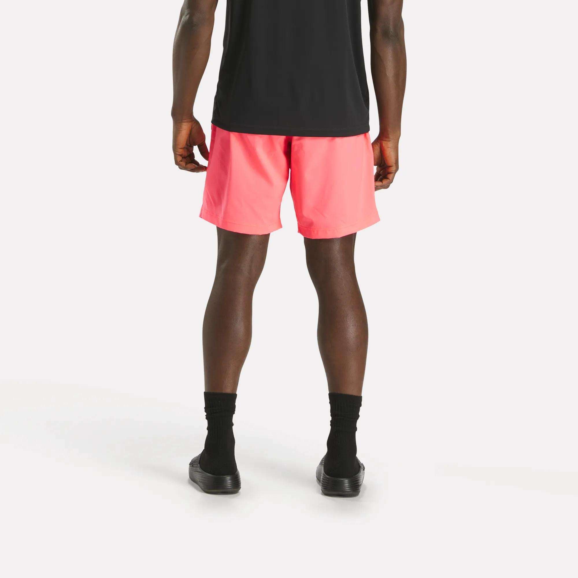 A person faces away from the camera, wearing a black shirt, black socks and shoes, and bright pink Reebok Men's Workout Ready Woven Shorts against a plain white background.