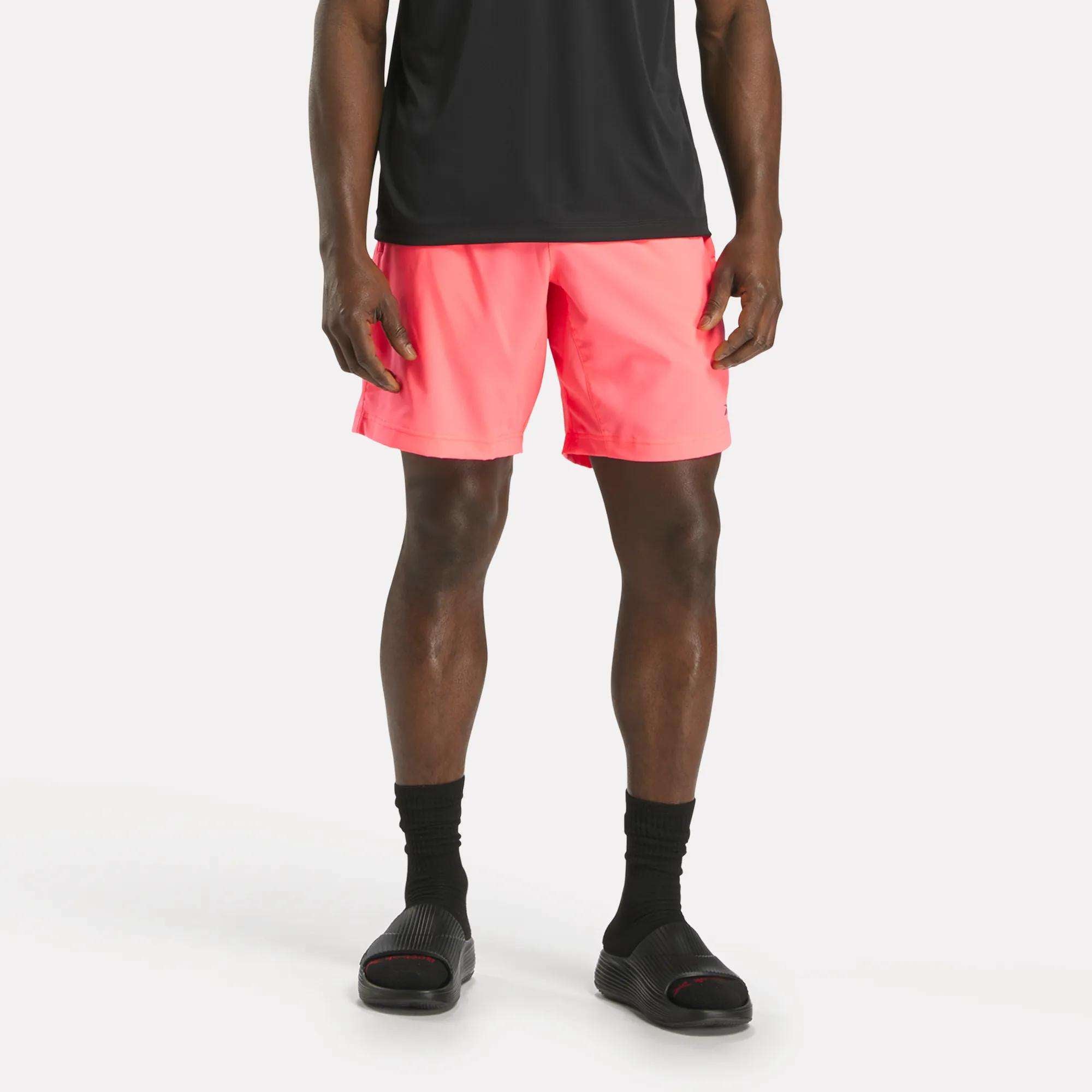 A person wearing a black t-shirt, Reebok Men's Workout Ready Woven Shorts in bright pink, black socks, and black slide sandals stands against a plain white background.
