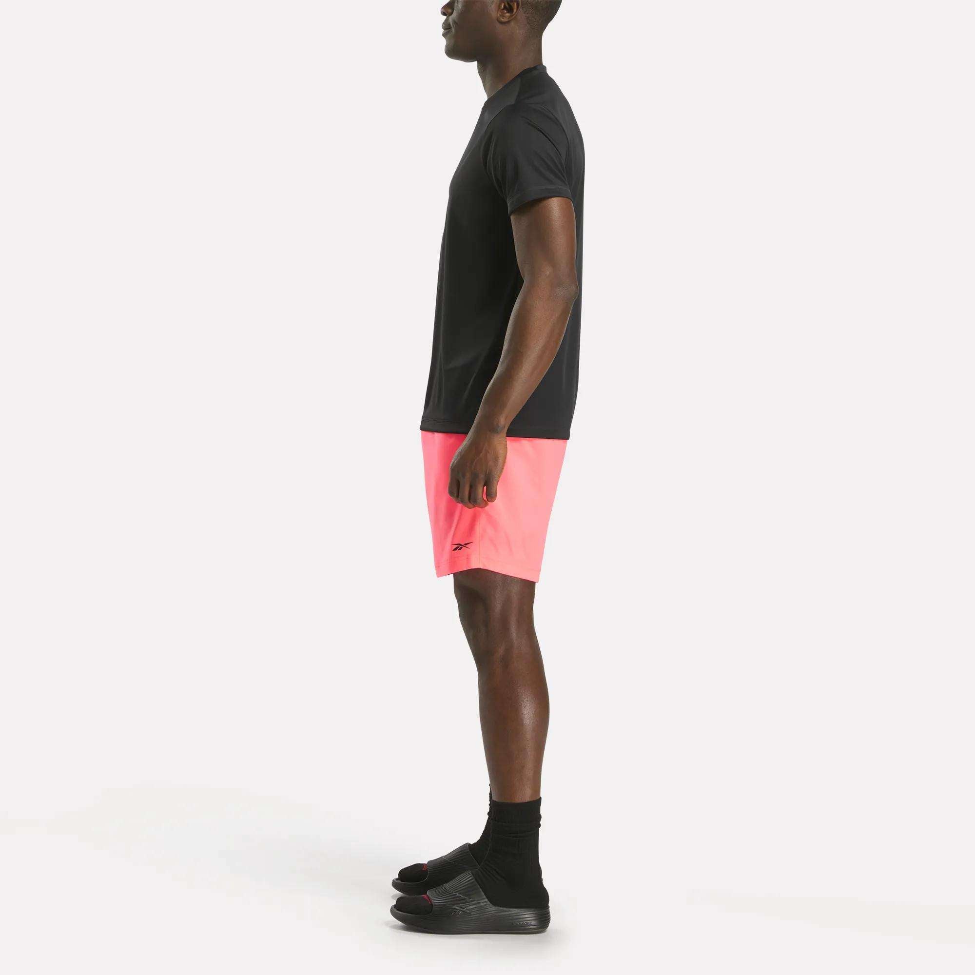 A person stands in profile against a plain white background wearing a black t-shirt, black socks, black slide sandals, and bright pink Reebok Men's Workout Ready Woven Shorts.