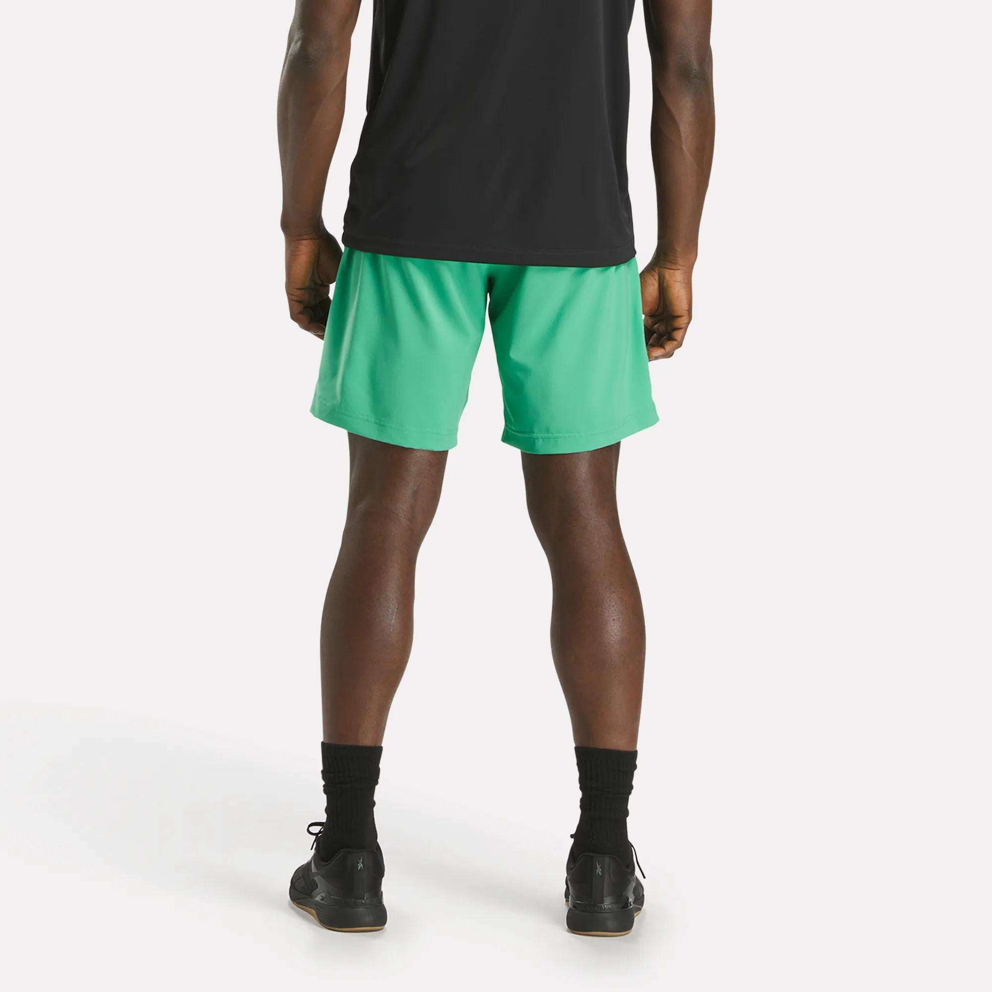 A person faces away from the camera, wearing a black shirt, black socks, black sneakers, and bright green Reebok Men's Workout Ready Woven Shorts against a plain white background.
