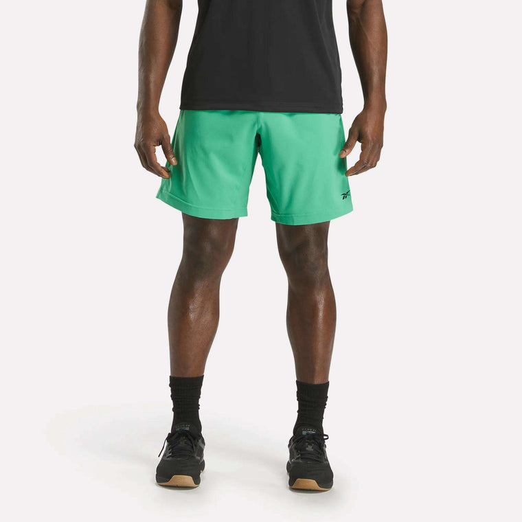 A person wearing a black t-shirt, Reebok Men's Workout Ready Woven Shorts in green, black socks, and black sneakers stands facing forward against a plain white background.