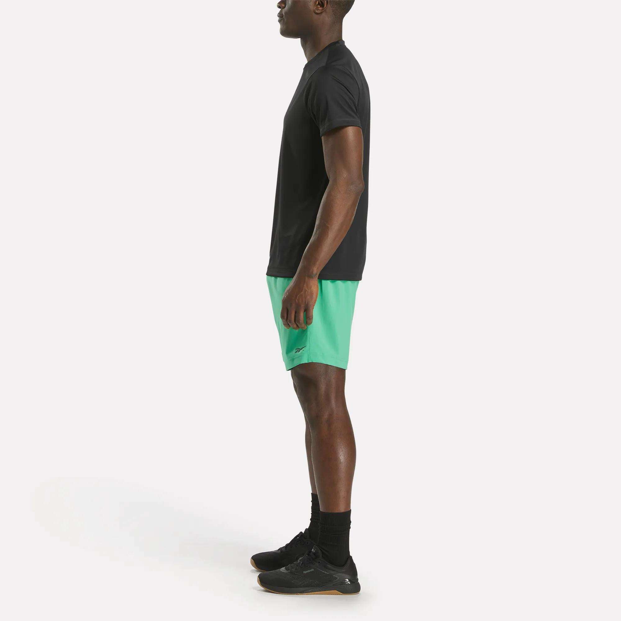 A man stands in profile view against a plain white background, wearing a black t-shirt, black socks, black sneakers, and green Reebok Men's Workout Ready Woven Shorts.
