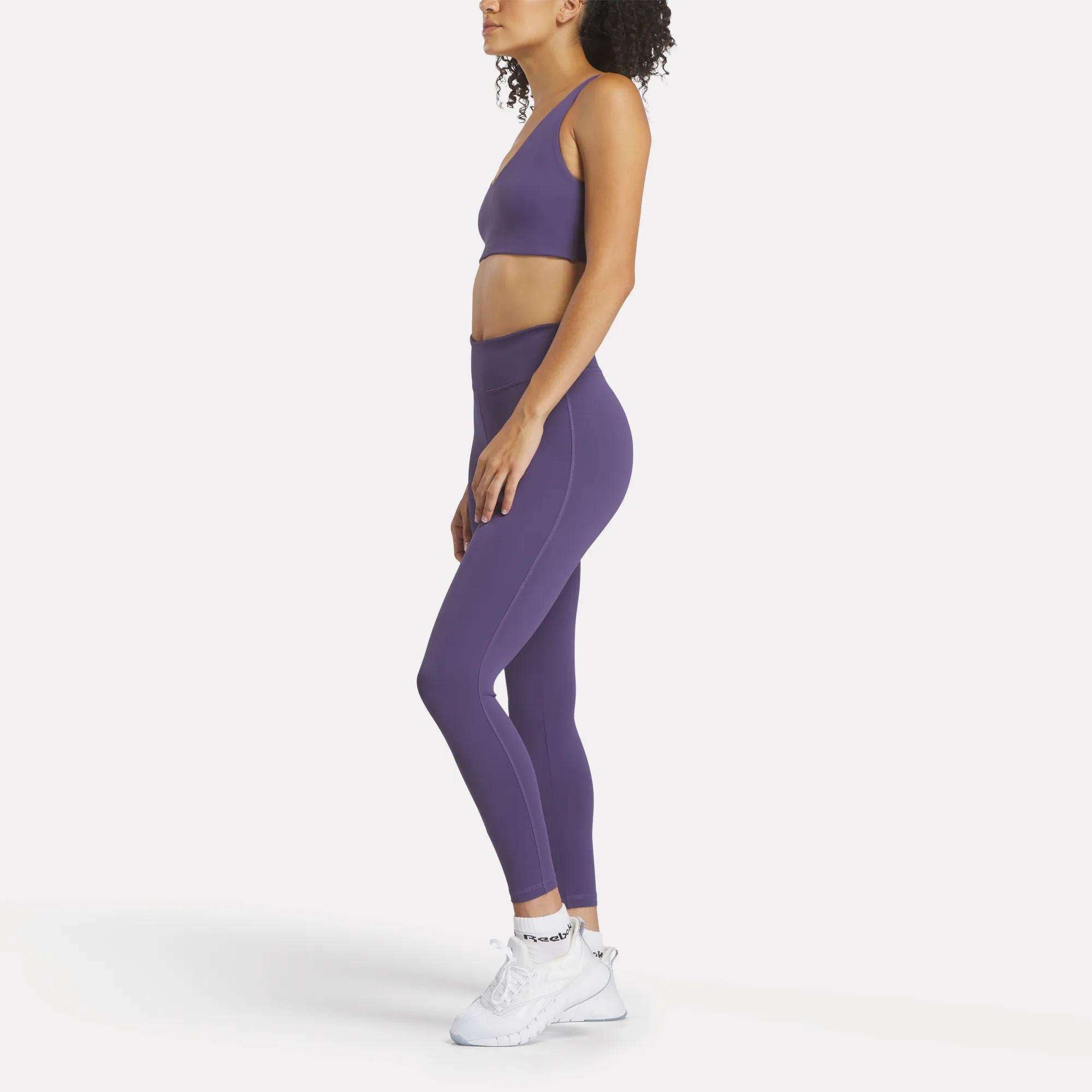 A woman stands in profile against a white background, wearing Reebok Women's Lux High-Rise Tights made from RBK-DRY performance fabric, a purple sports bra, and women's Reebok athletic shoes.