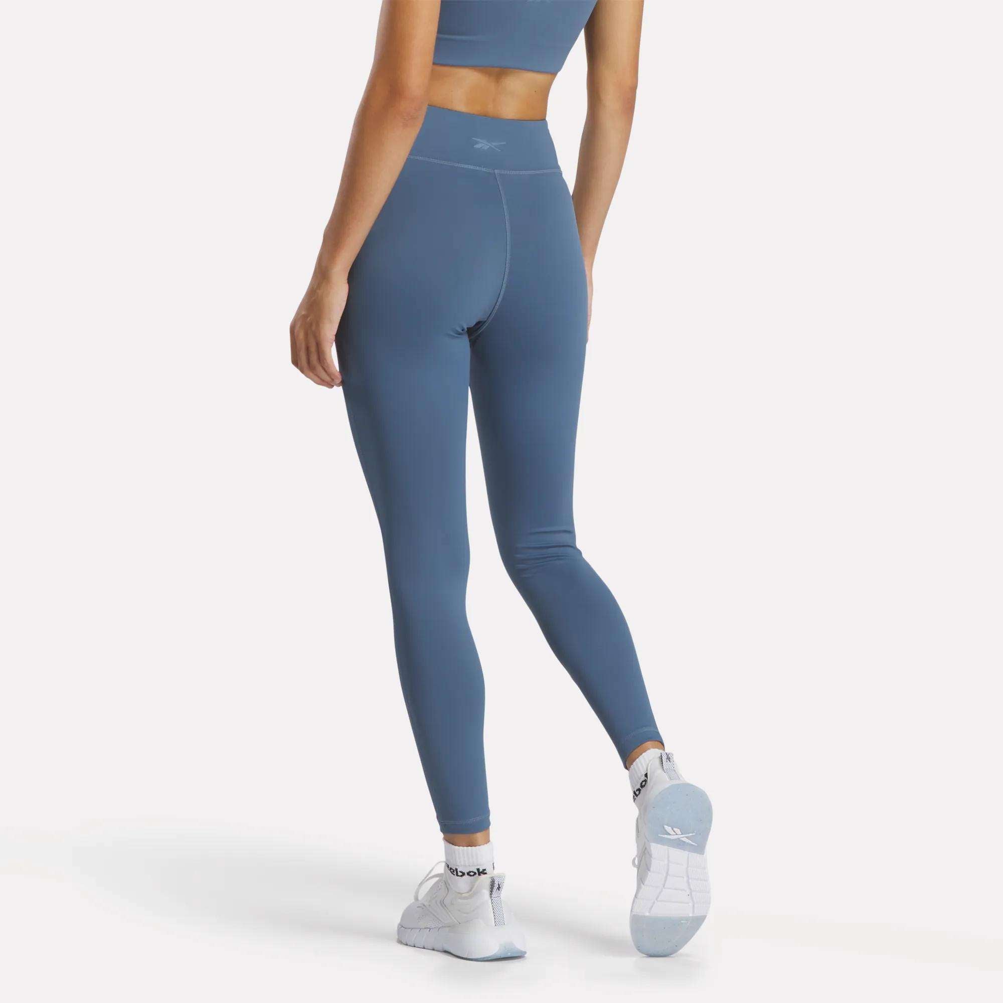 A woman stands on one leg facing away, wearing a blue Reebok sports bra and Women's Lux High-Rise Tights, both in RBK-DRY fabric, paired with white athletic socks and white sneakers against a plain white background.