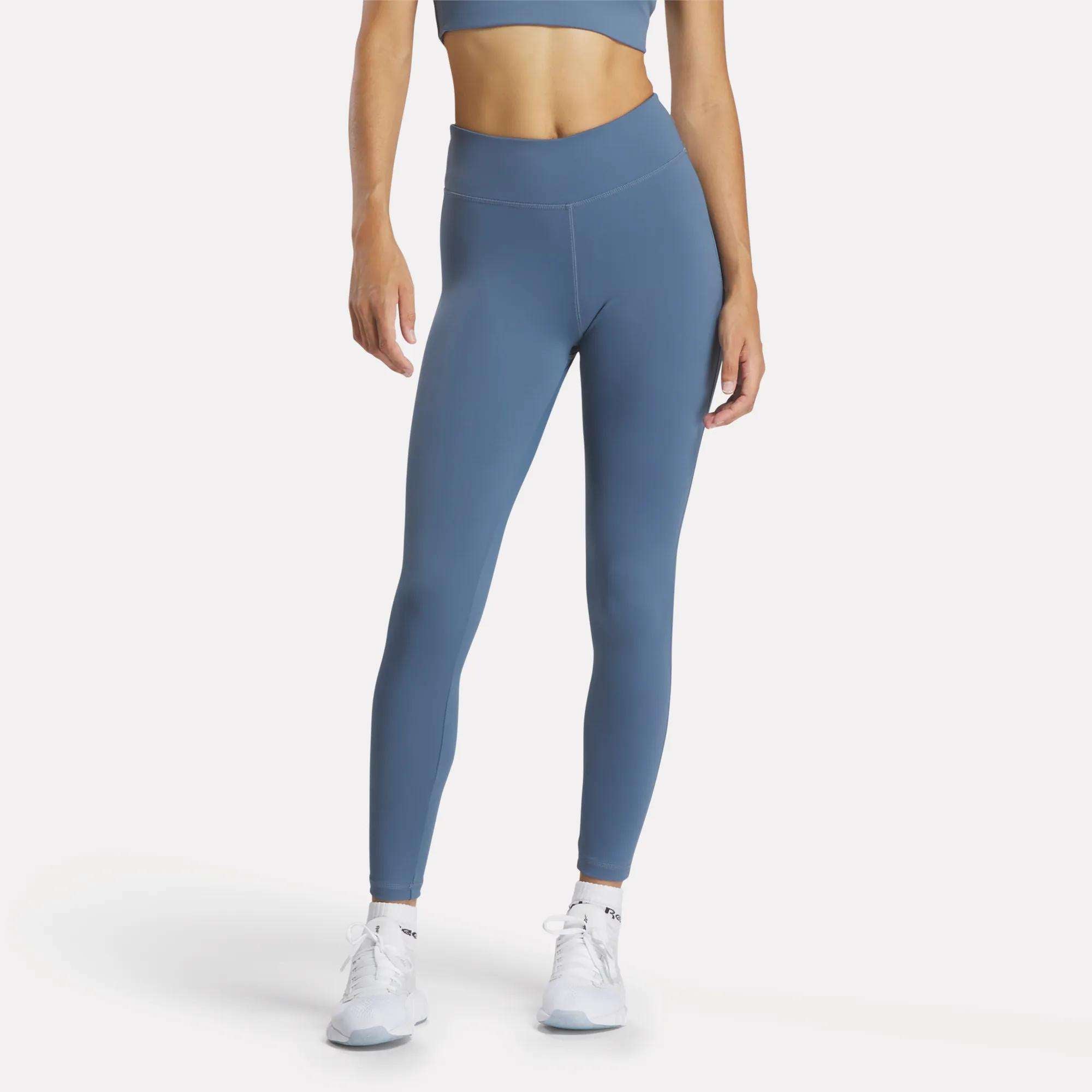 A person wears a blue Reebok sports bra and Women's Lux High-Rise Tights, made with RBK-DRY performance fabric, standing against a plain white background with white athletic shoes.