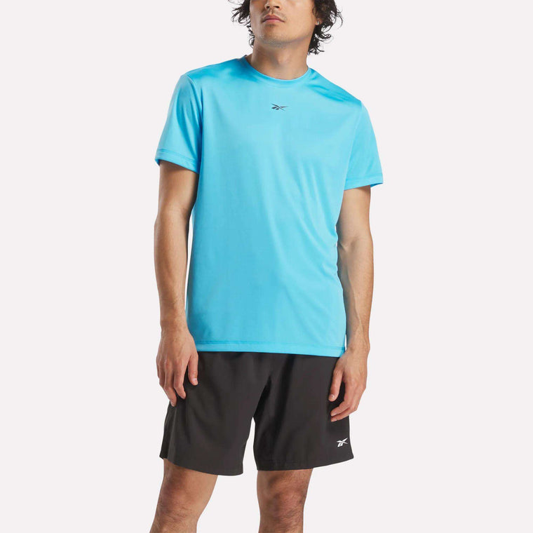 A person wearing a bright blue Reebok Men's ID Train Short Sleeve Tech Tee and black shorts with a small Reebok logo stands against a plain light background.