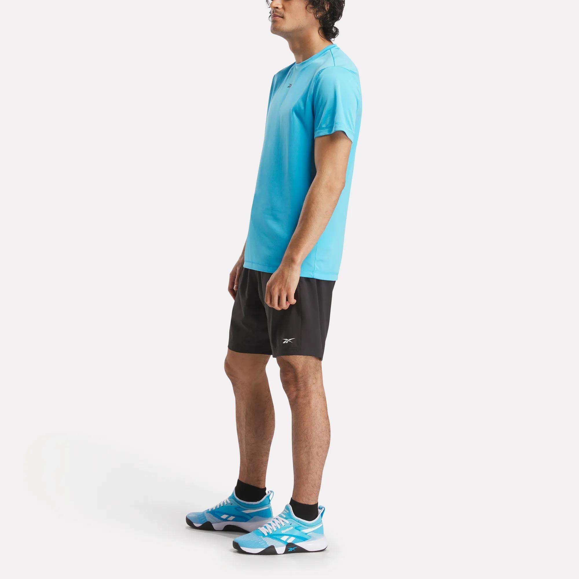 A man stands in profile on a white background, wearing a bright blue Reebok Men's ID Train Short Sleeve Tech Tee, black shorts, black socks, and blue athletic shoes.