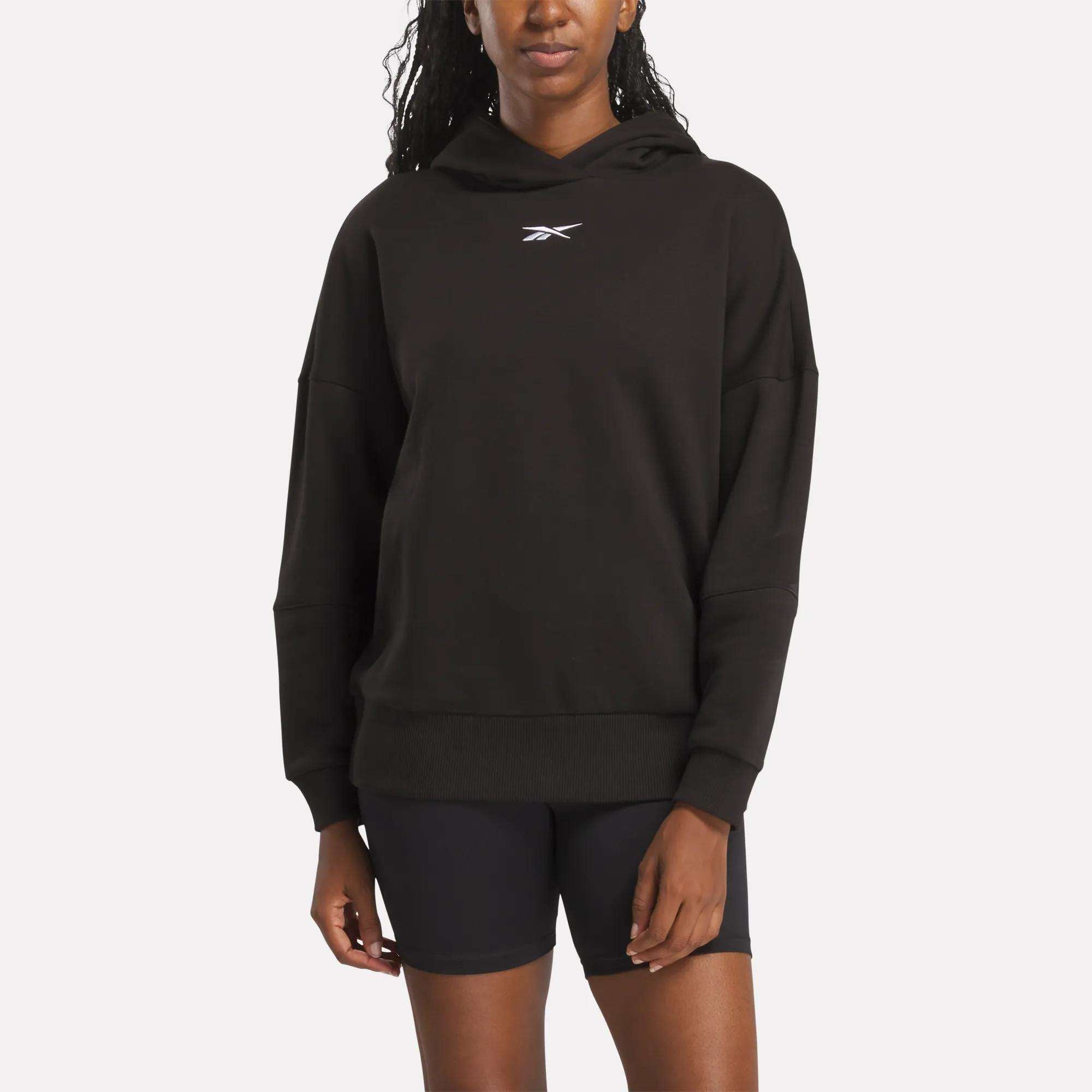 A person wears the Reebok Women's Lux Hoodie with a small white logo on the chest and black shorts, standing against a plain light background.
