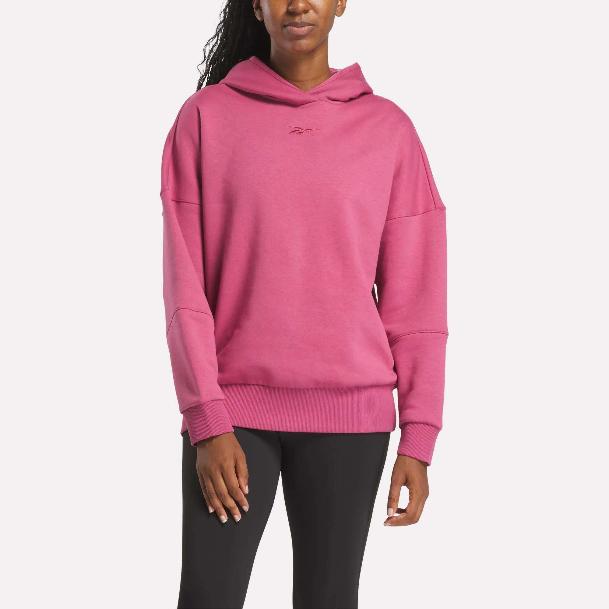 A woman stands against a plain white background wearing the Reebok Women's Lux Hoodie in pink, styled loosely and paired with black leggings.
