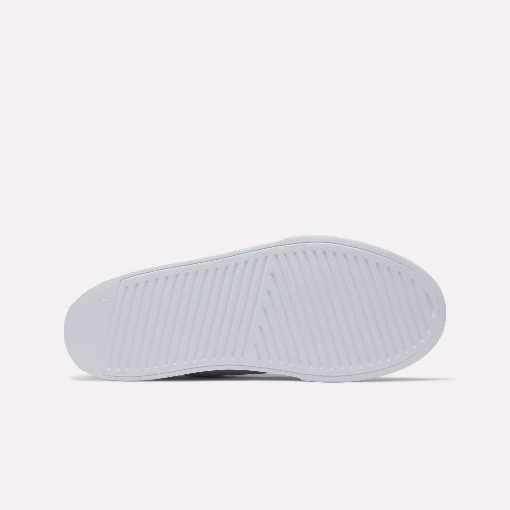 The image shows the sole of a white Women's OnLux Court Lite Walking Shoe by Reebok, viewed from the bottom. The sole has a textured pattern with diagonal and crosswise grooves for traction. The background is plain and light-colored.
