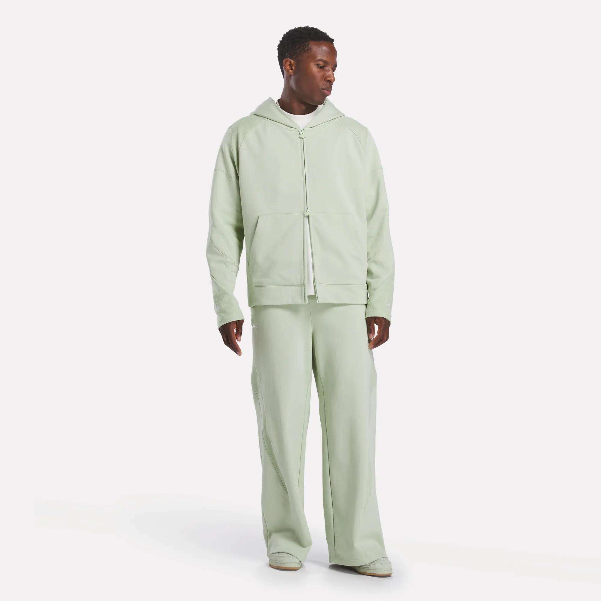 A man wears a light green Reebok x CHUKWU Full-Zip Hoodie and matching loose-fit pants, standing against a plain white background with white shoes, looking slightly down to his left.