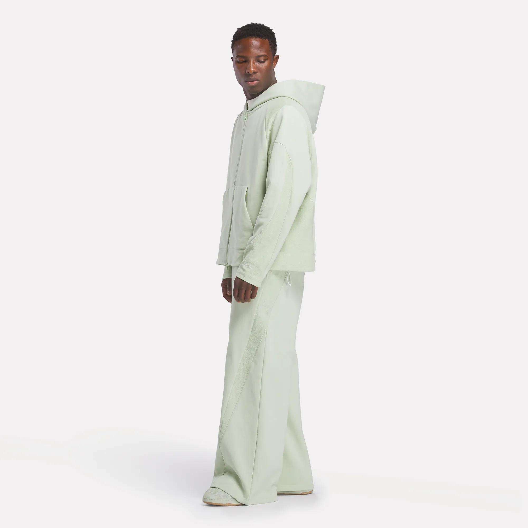A person stands against a plain white background, wearing the Reebok x CHUKWU Full-Zip Hoodie in light green with matching wide-leg pants and white shoes. They face slightly to the side with a relaxed posture. Brand: Reebok.