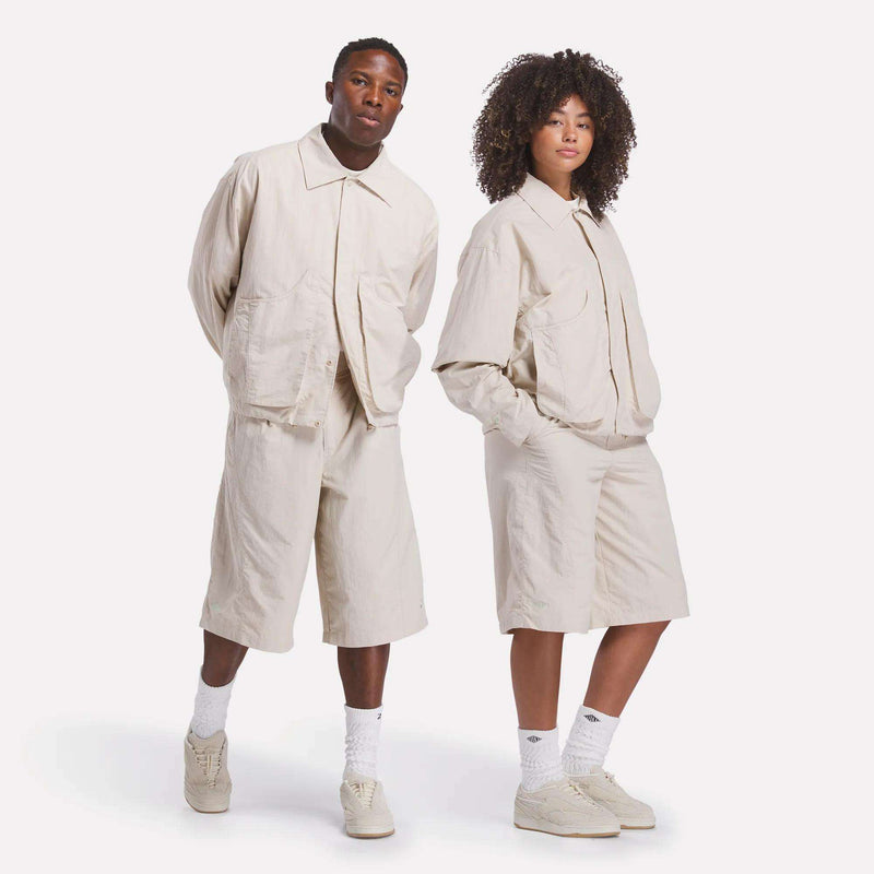 Two people stand side by side against a white background, both wearing matching light beige jackets and Reebok x CHUKWU High-Waist Shorts from Reebok, paired with white socks and beige sneakers.
