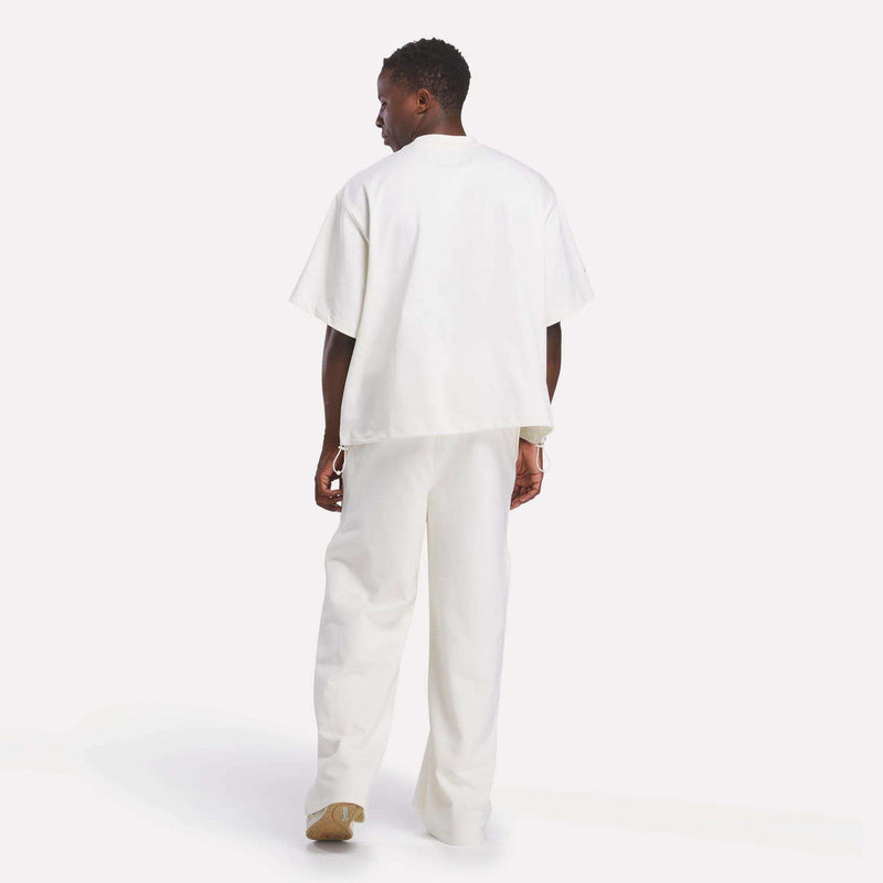 A man faces away, wearing an oversized white Reebok x CHUKWU Tee and loose white pants. The plain, light background has mint tones, and he pairs the look with soft light brown shoes.