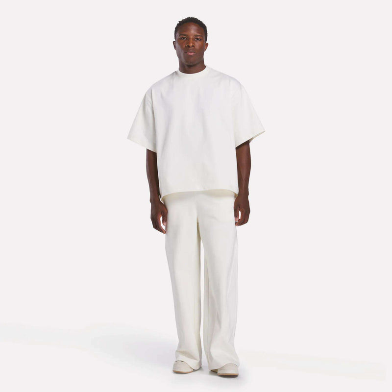 A man faces forward wearing a loose Reebok x CHUKWU Tee by Reebok, paired with wide white pants and shoes on a plain white background, channeling Tobe Nwigwe’s signature style with subtle mint accents.