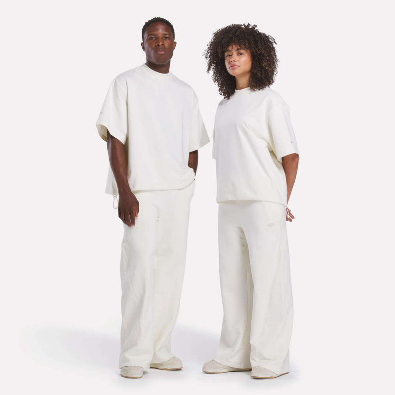 Two people stand side by side against a plain white background wearing loose-fitting, off-white Reebok x CHUKWU Tees and wide-leg pants. One top is subtly accented with mint tones. Both face forward.