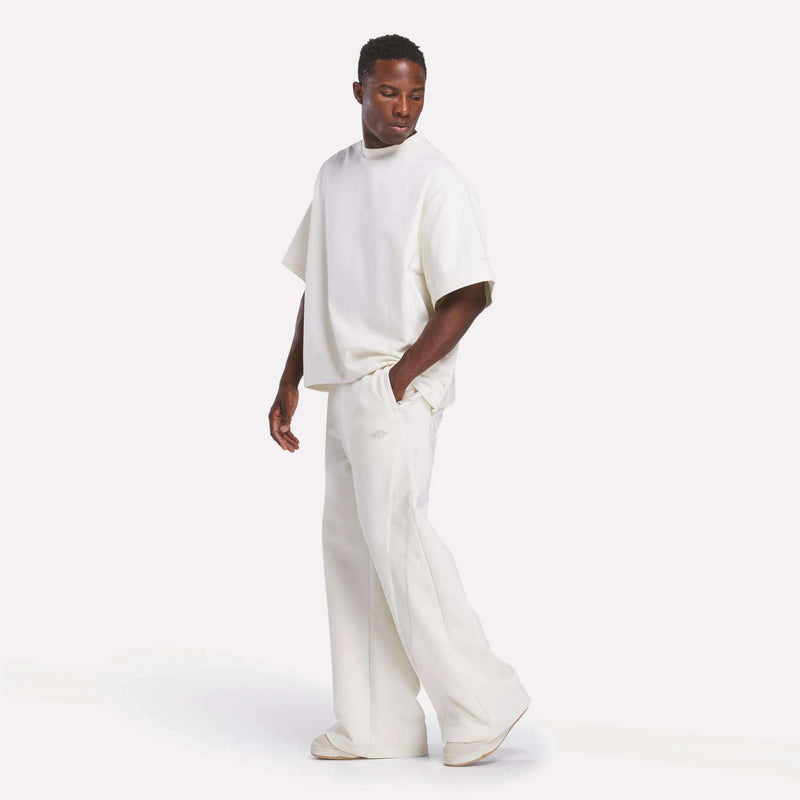A man stands against a plain white background, wearing a loose-fitting Reebok x CHUKWU Tee and wide-leg white pants with matching shoes, looking down to his left in a minimalist style reminiscent of Tobe Nwigwe.
